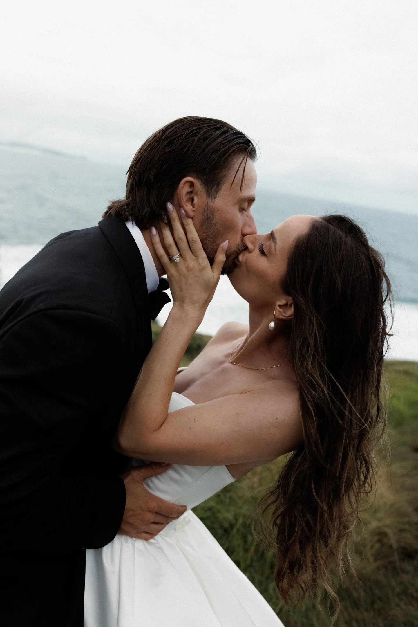 A newlywed couple sharing a kiss outdoors near the ocean, the groom in a black tuxedo and the bride in a strapless white wedding gown with long wavy hair.