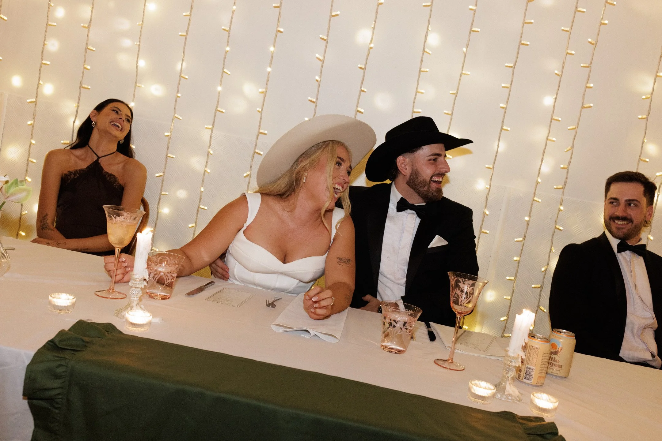 A group of people dressed in formal attire, sitting at a decorated table with candles, glasses, and tableware, enjoying a celebration in a festively lit setting.