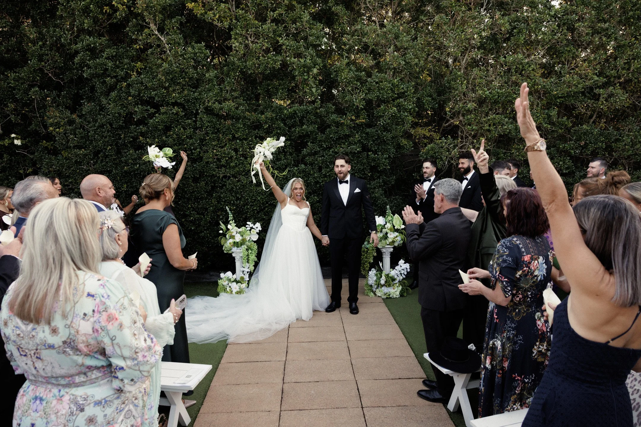 Bride and groom at a wedding ceremony outdoors, surrounded by friends and family, with the bride holding a bouquet of white flowers, and guests clapping and celebrating.