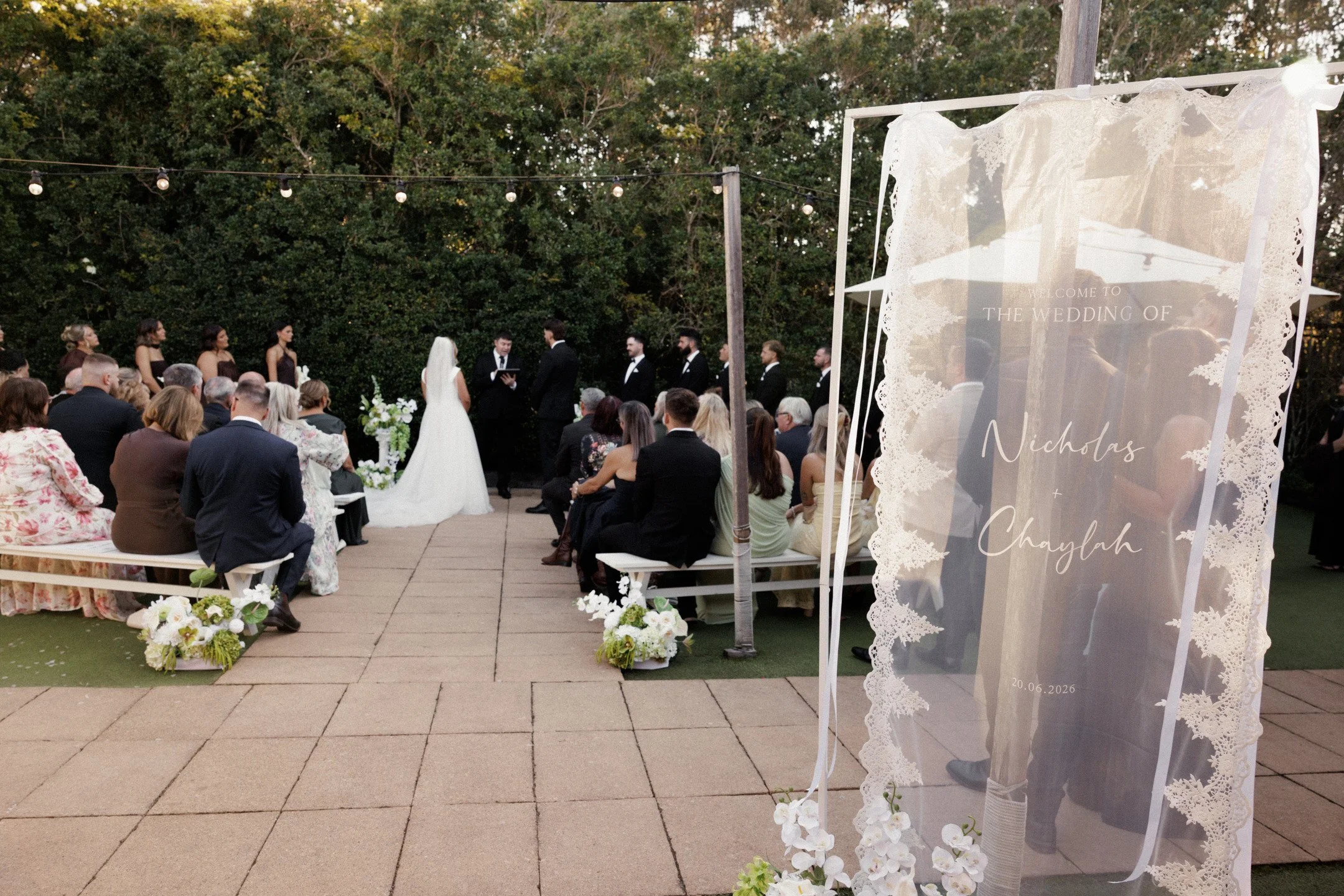 Outdoor wedding ceremony with guests seated, bride and groom standing under arch, surrounded by trees, decorative white sign with floral accents reading 'Welcome to the wedding of Nichols and Chaylah' outdoors.