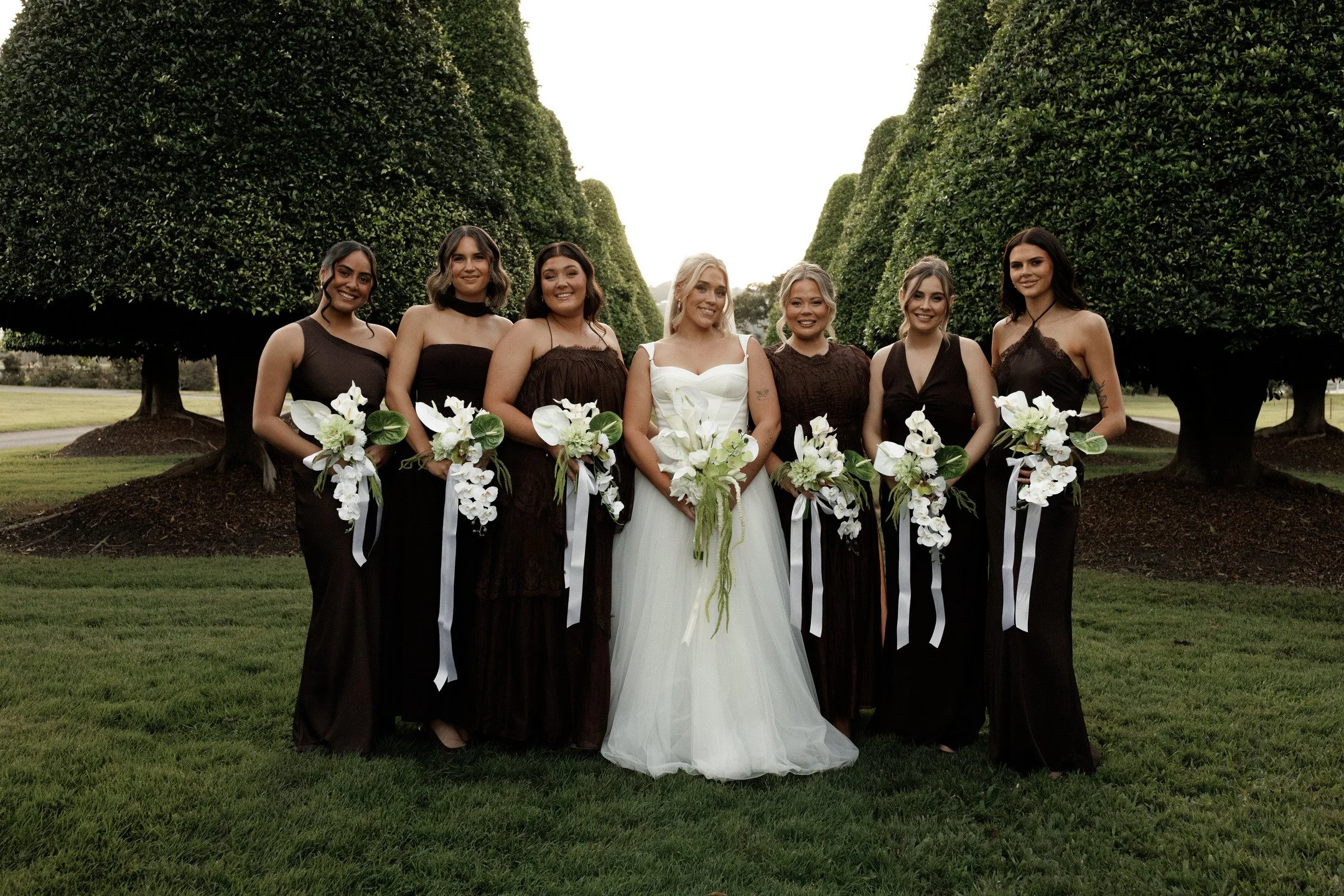 Group of seven women at a wedding outdoors, wearing formal dresses and holding bouquets, standing on grass with green, trimmed trees in the background.