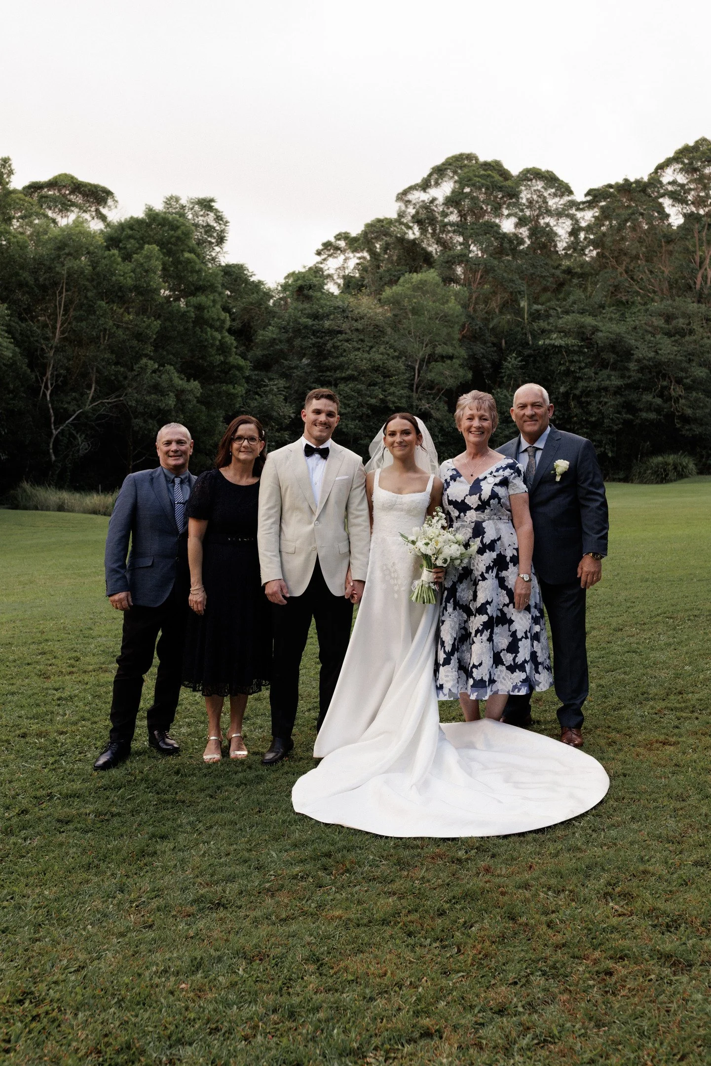 A wedding photo of six people, including a bride and groom, standing on a grassy field with trees in the background. The bride is in a white wedding gown holding a bouquet, and the groom is in a white tuxedo. The others are family members dressed in 