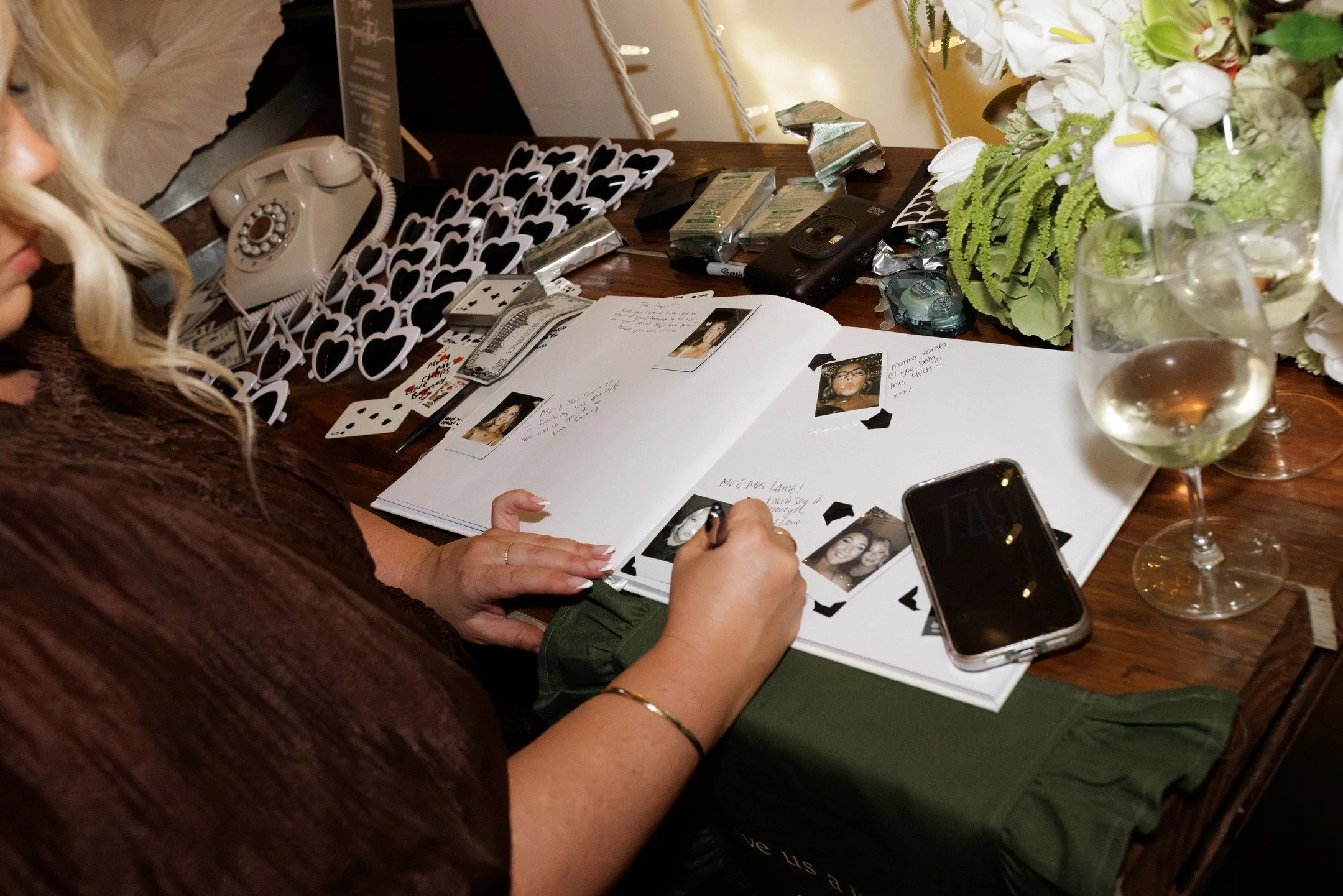A woman with long, curly blonde hair is looking at a guestbook or photo album on a wooden table, surrounded by black and white heart-shaped photo props, playing cards, a vintage rotary phone, and several gift-wrapped items. To the right, there are tw