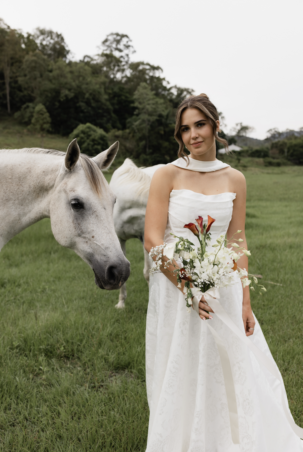 A young woman in a white wedding dress holding a bouquet of flowers standing in a green field next to a white horse.