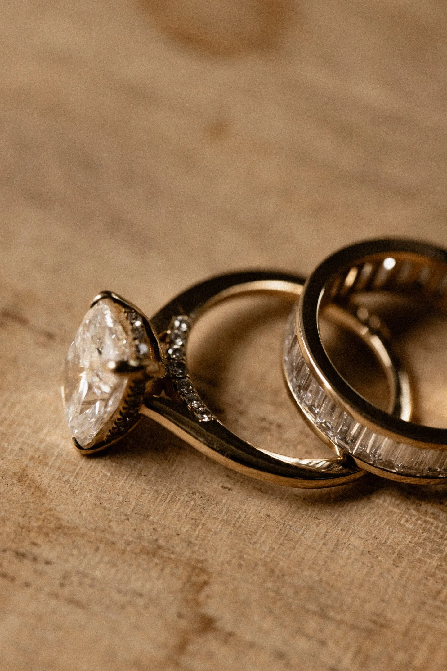 Close-up of two rings on a wooden surface, one with a large central gemstone and smaller stones surrounding it, the other with a band of small rectangular stones.