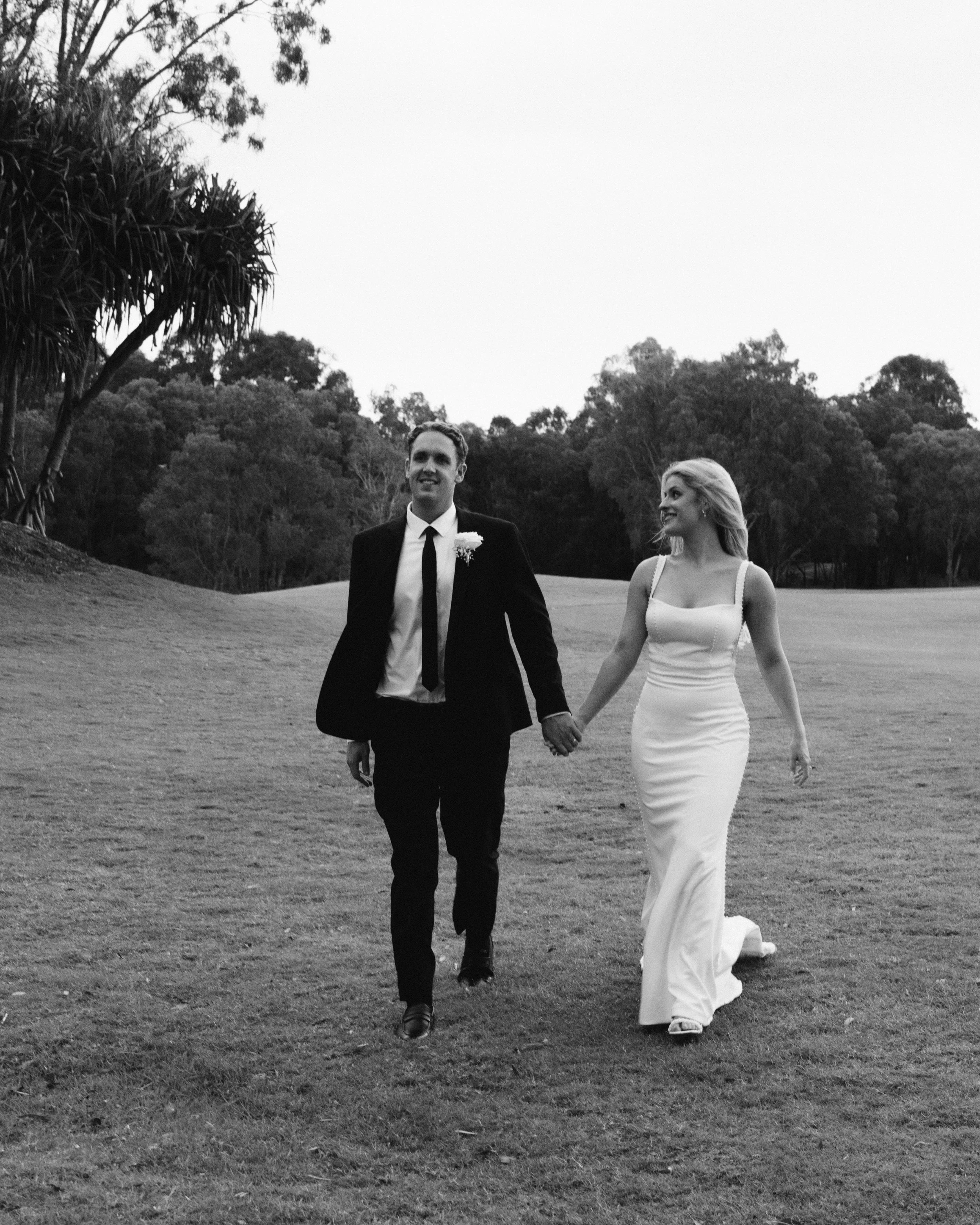 Black and white photo of a bride and groom walking hand in hand outdoors, surrounded by trees and grass.
