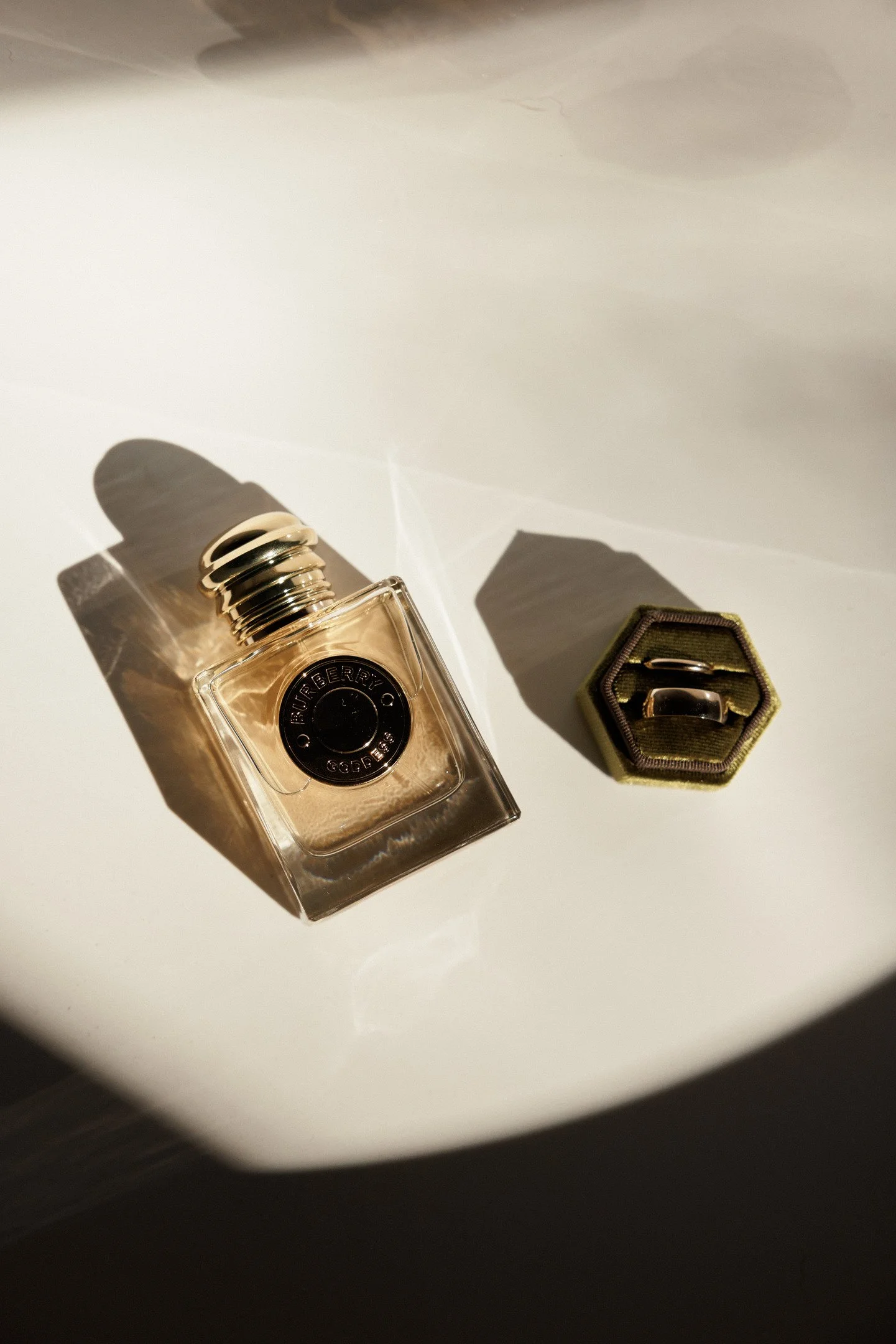 A partially visible Burberry perfume bottle with a black circular label and a metallic cap, reflecting light and casting shadows, and a small hexagonal box with a pair of rings inside, on a white surface.
