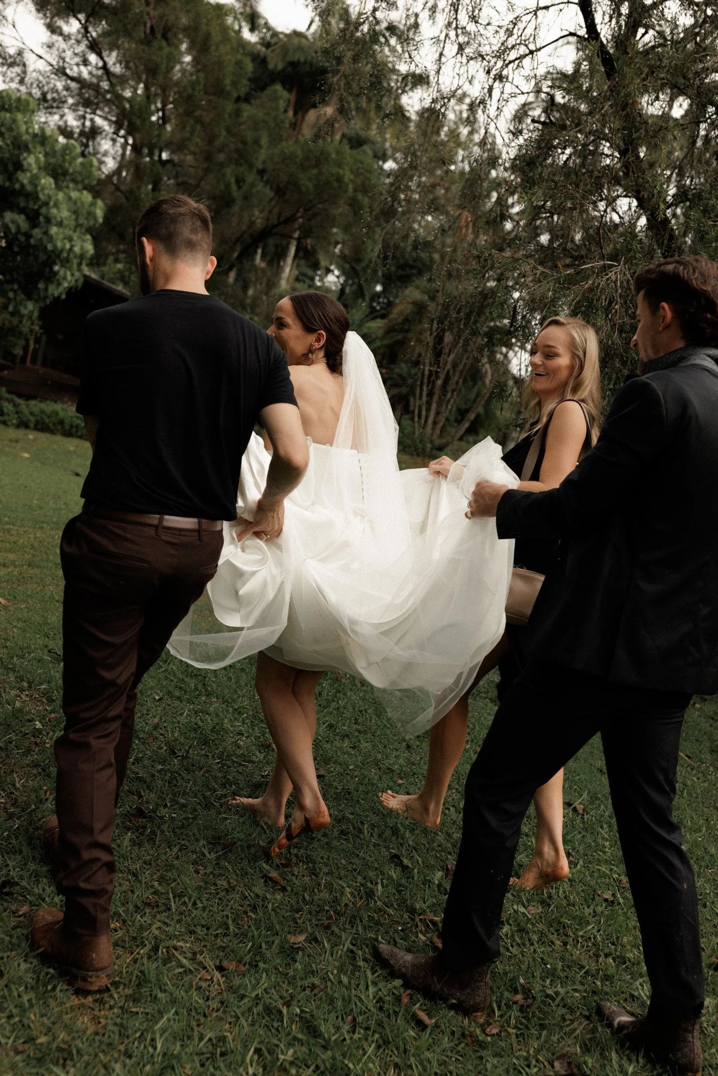 Four people, dressed in formal attire, are outdoors in a grassy area surrounded by trees. They are holding and lifting a woman dressed in a wedding gown and veil, all smiling and barefoot.