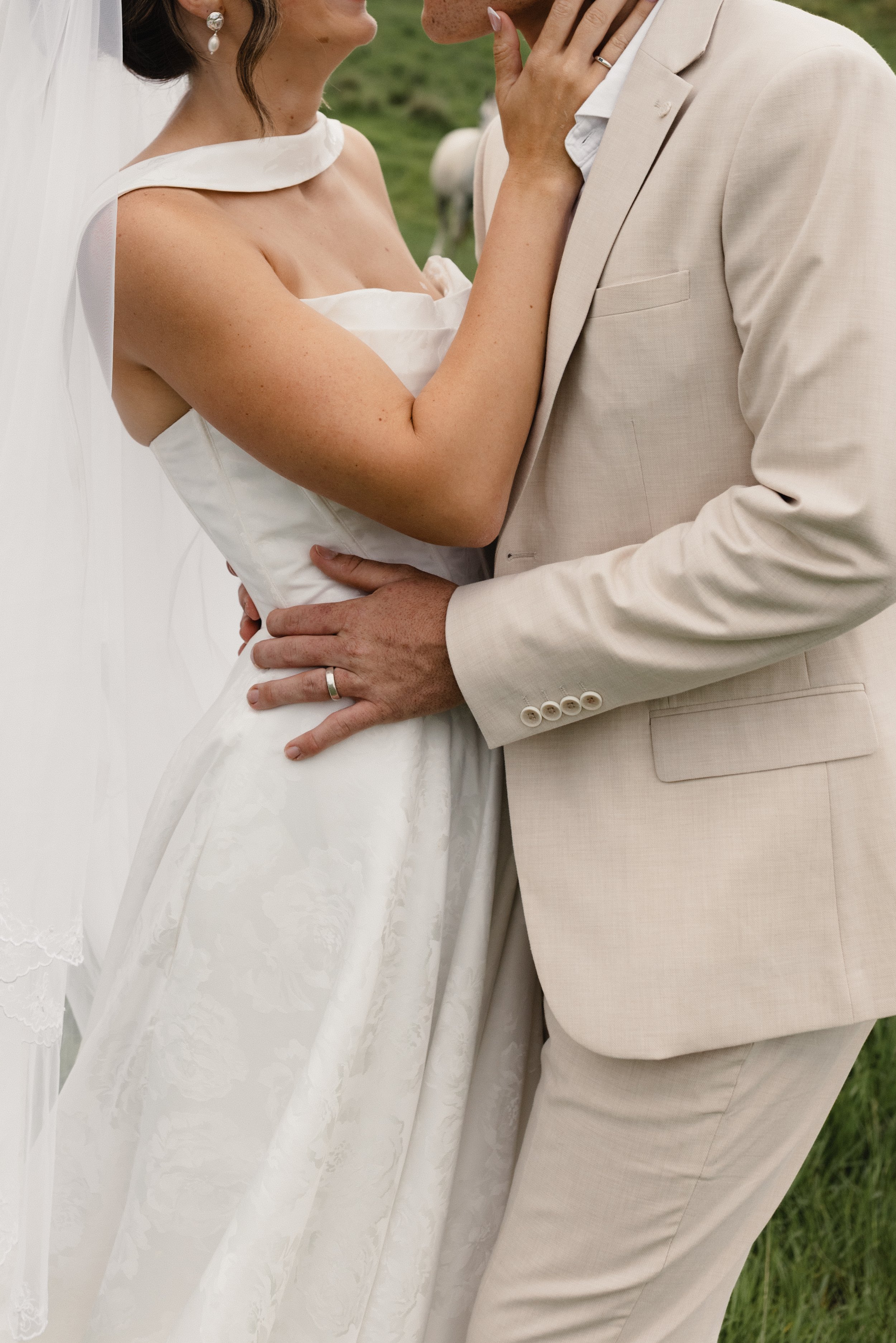 Close-up of a bride and groom in wedding attire, touching foreheads and holding each other, outdoors on grass.