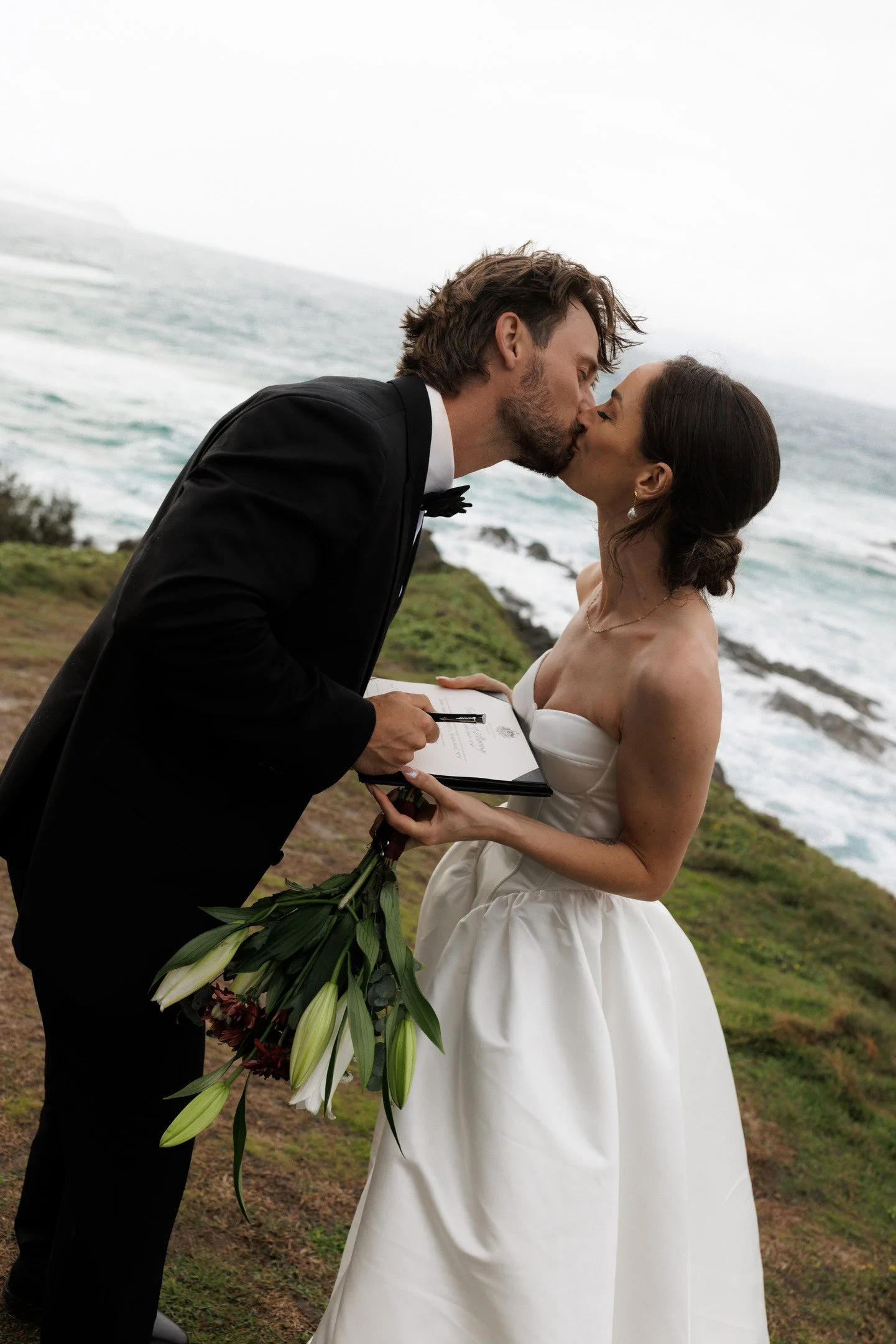 A couple getting married outdoors by the ocean, kissing during their wedding ceremony. The groom is in a black tuxedo holding a bouquet of flowers, and the bride is in a white strapless wedding dress holding an open book and a bouquet of lilies.