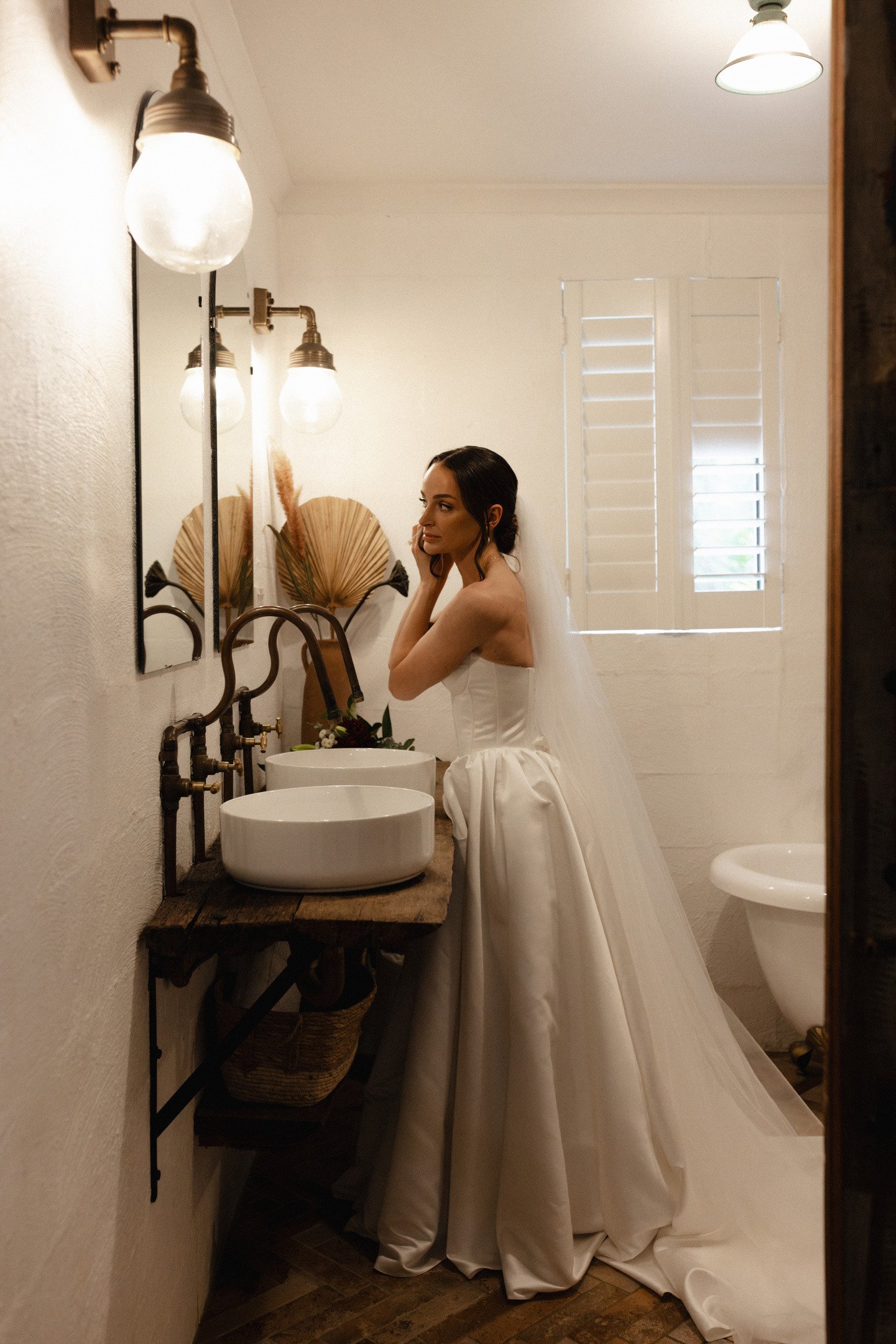 A woman in a wedding gown inside a rustic-style bathroom, looking at herself in a mirror above a wooden countertop with farmhouse sinks and vintage-style lighting. The bathroom has white walls, shuttered window, and decorative dried palm leaves on th