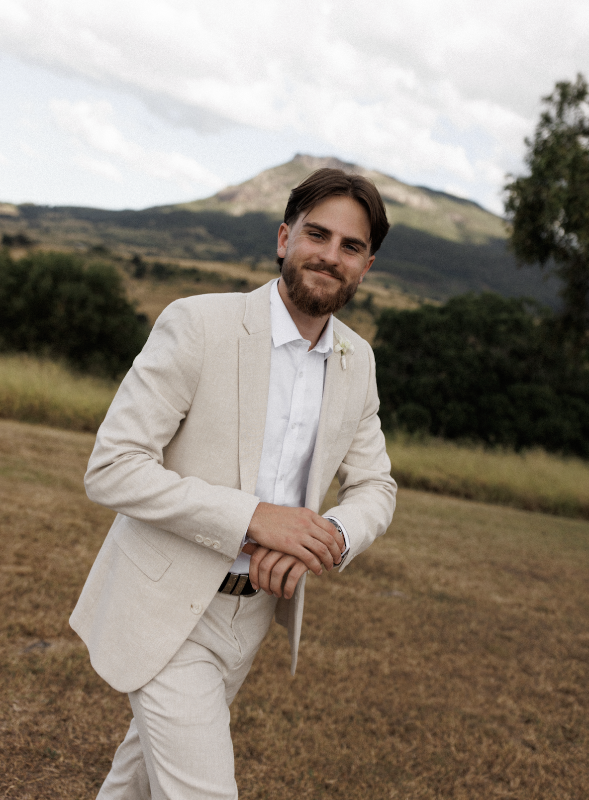 A young man in a cream-colored suit and white shirt outdoors with mountains and trees in the background.
