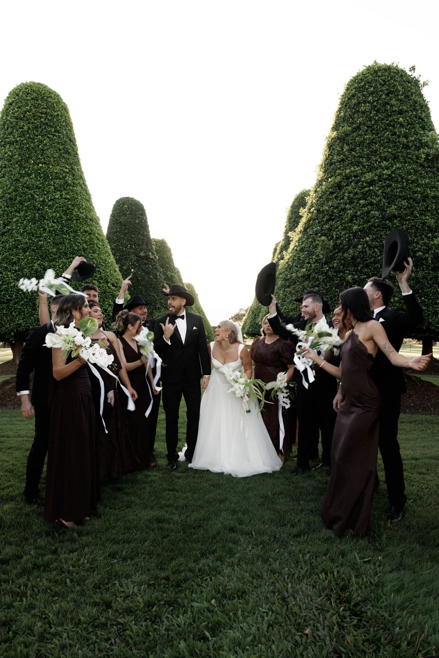 A wedding celebration outdoors with a bride in a white gown and a groom in a black tuxedo, surrounded by friends in formal attire, holding hats and flowers, with tall, neatly trimmed green trees lining the scene.
