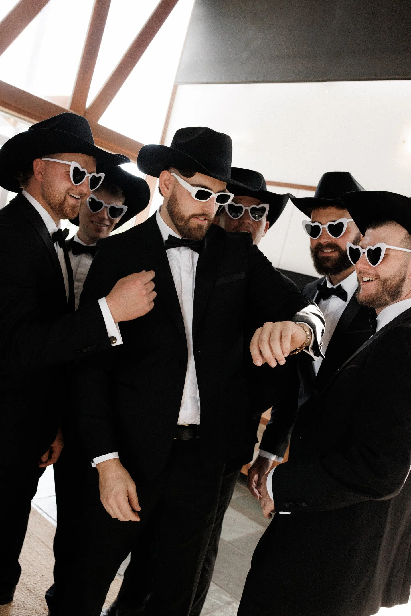 A group of six men dressed in black tuxedos, wearing black hats and white heart-shaped sunglasses, gathered together and looking at a watch one of them is wearing.