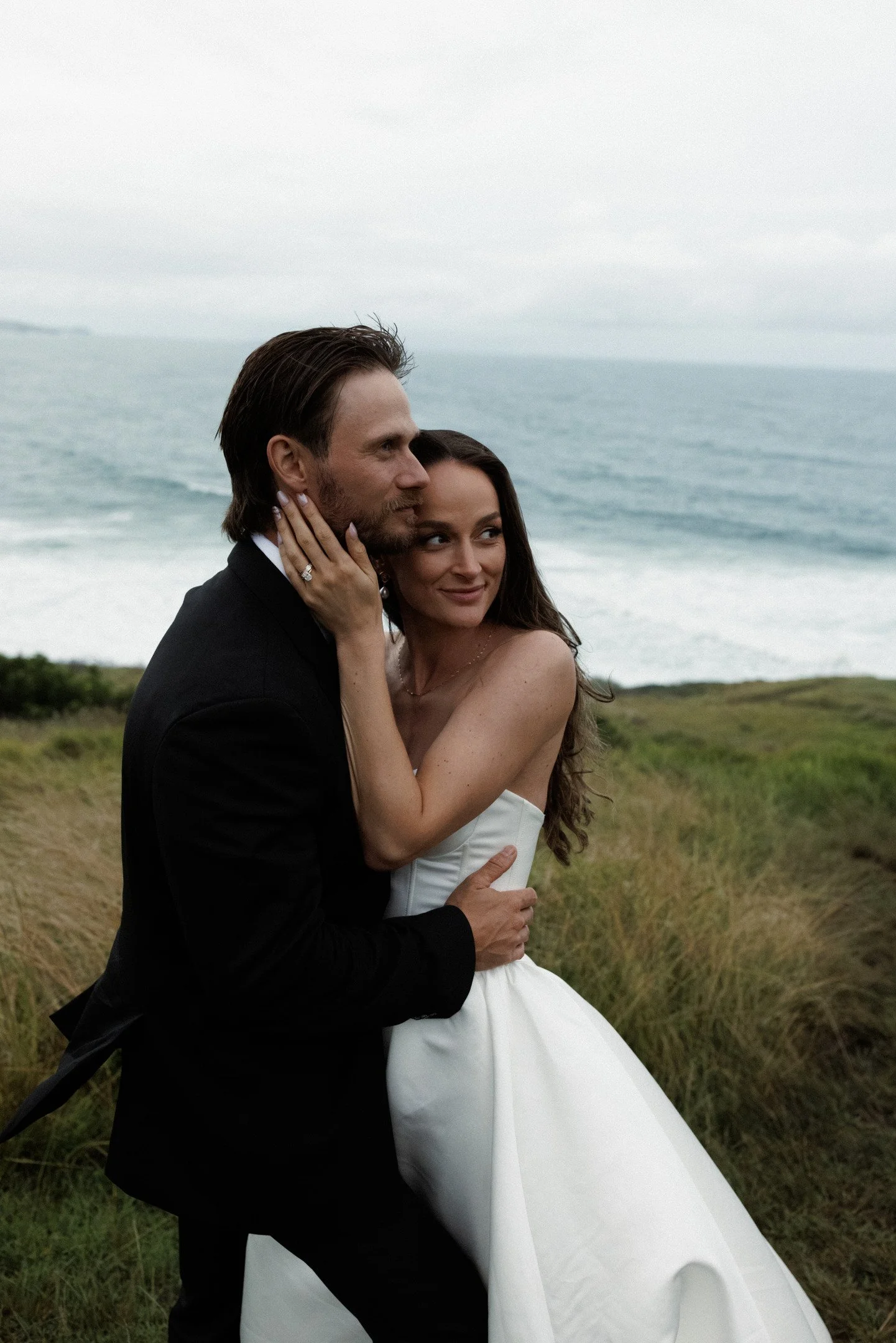 A bride and groom embrace outdoors by the ocean, with grassy dunes and cloudy sky in the background.