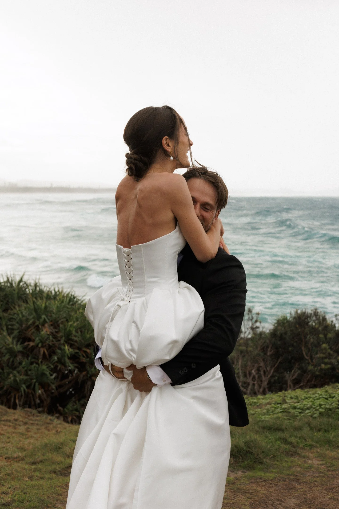 A bride and groom embrace on a beachside with ocean waves and overcast sky in the background.