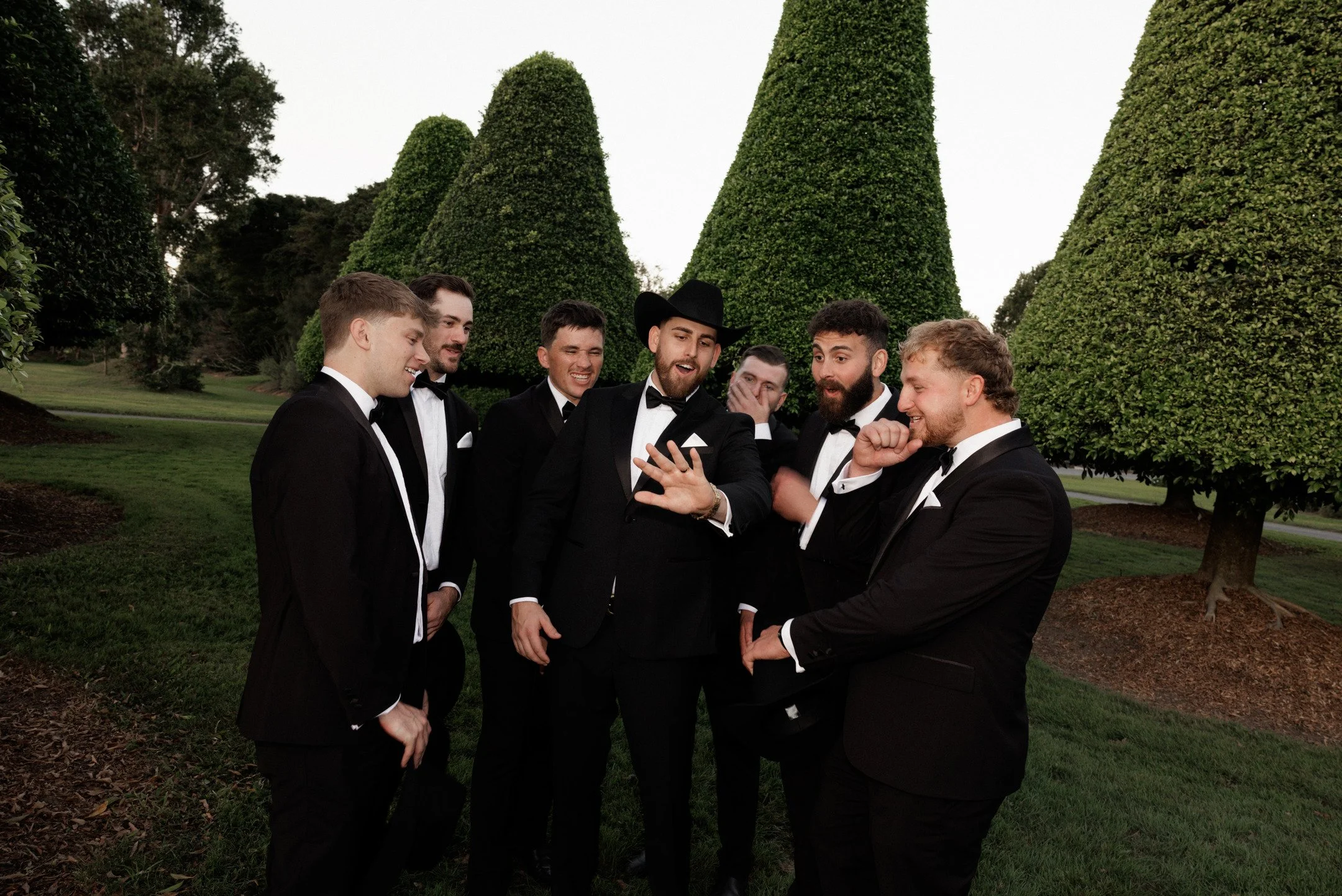 Group of seven men in tuxedos and one man in a cowboy hat and tuxedo, smiling and talking outdoors with large green trees in the background.