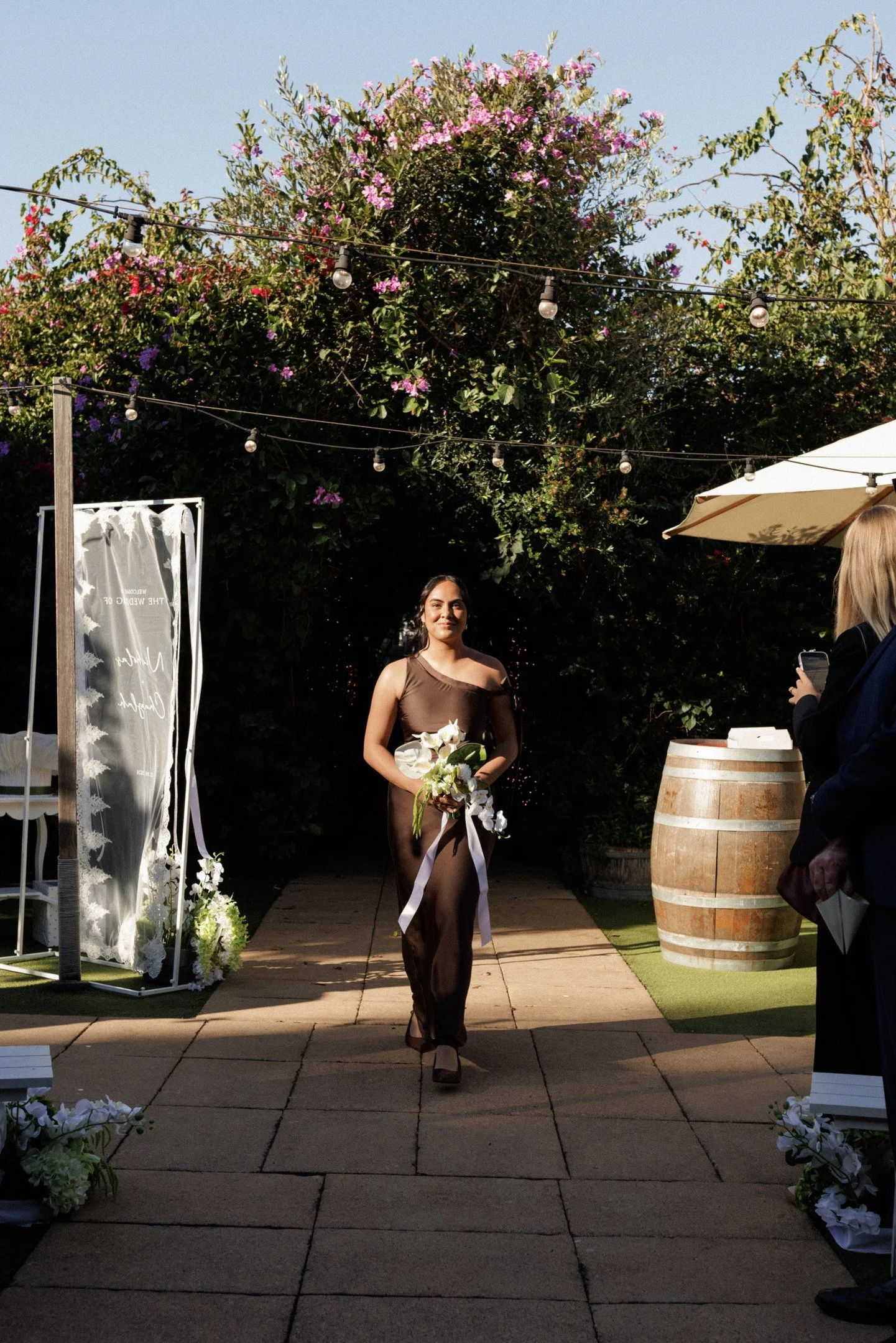 A woman in a sleeveless dark brown dress holding a bouquet of white flowers walks along an outdoor aisle decorated with flowers and greenery. There are string lights overhead, a white canopy, and a large barrel on the right. A person with blonde hair