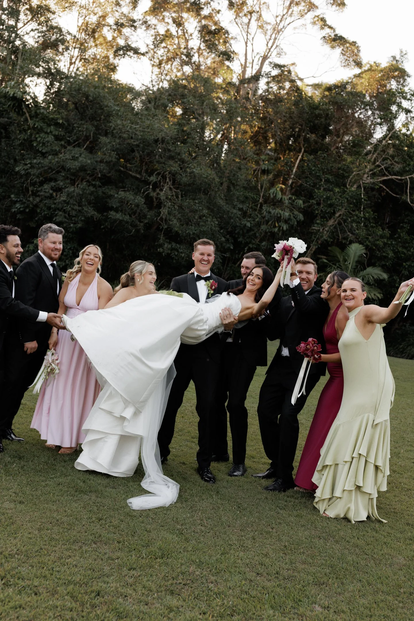 A wedding party outdoors, with the groom carrying the bride, surrounded by friends and family, during sunset in a lush green garden.
