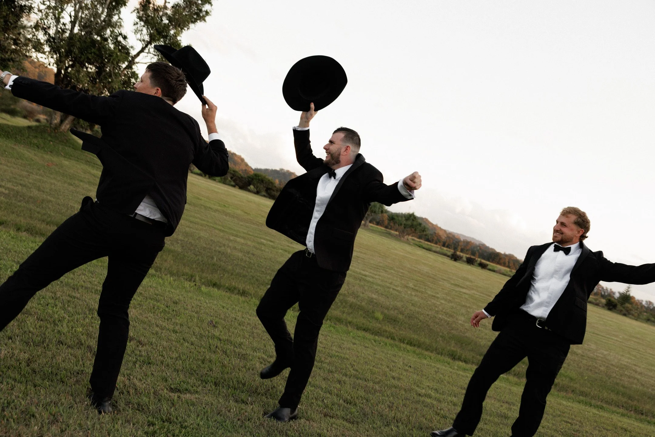 Three men in formal tuxedos and bow ties are outdoors on a grassy field, celebrating and tossing black hats into the air, with trees and cloudy sky in the background.