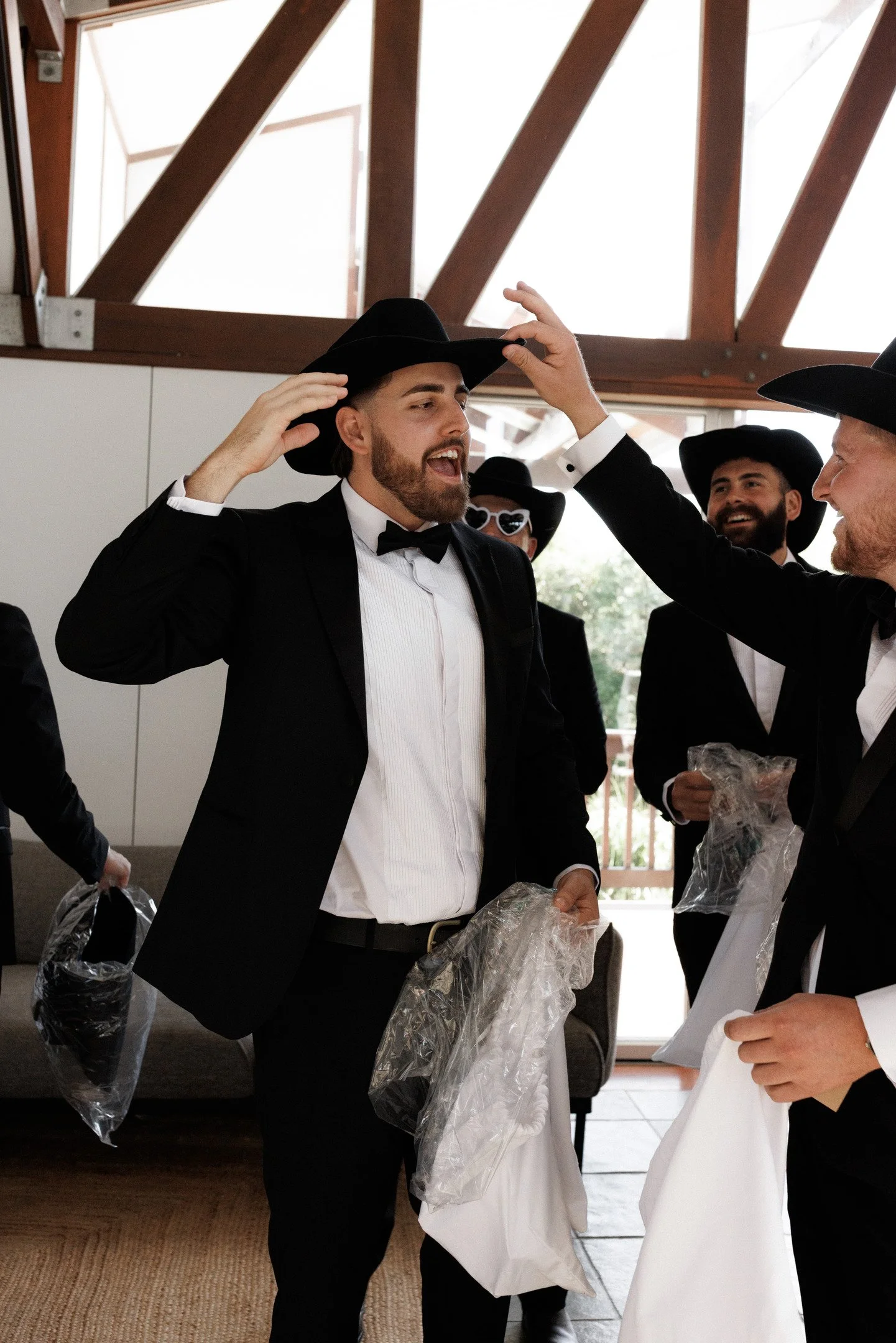 Group of men dressed in formal tuxedos and black hats, gathered indoors, engaged in conversation and smiling, with some holding plastic bags.