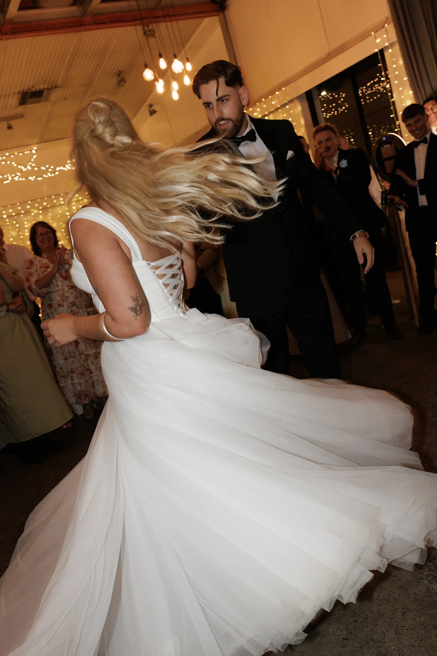 A bride dancing with a groom at a wedding reception, with guests smiling and watching in the background, decorated with fairy lights.