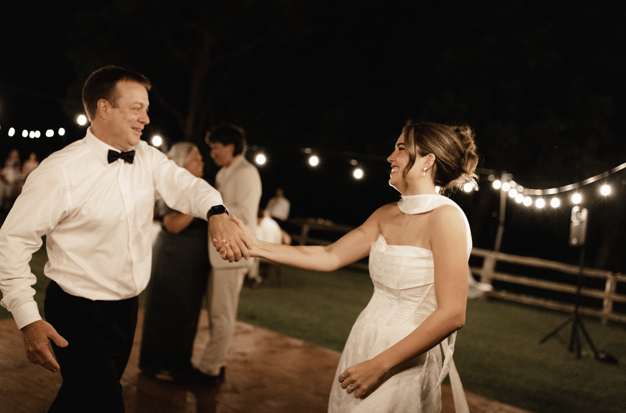 A bride in a wedding dress and an older man in a white shirt with black bow tie dancing and holding hands at night during a wedding reception. String lights illuminate the scene with several guests in the background.