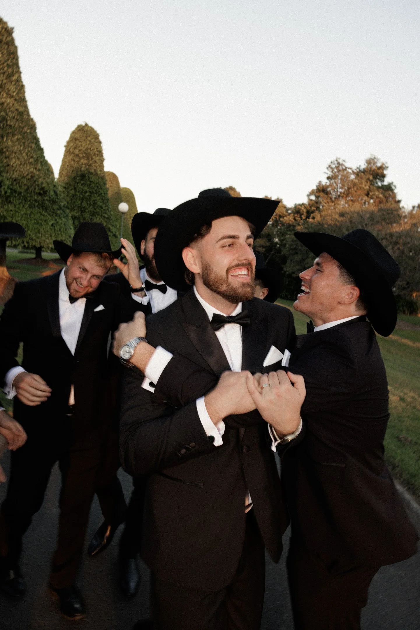Group of men in tuxedos and cowboy hats smiling and interacting outdoors during sunset, with trees in the background.