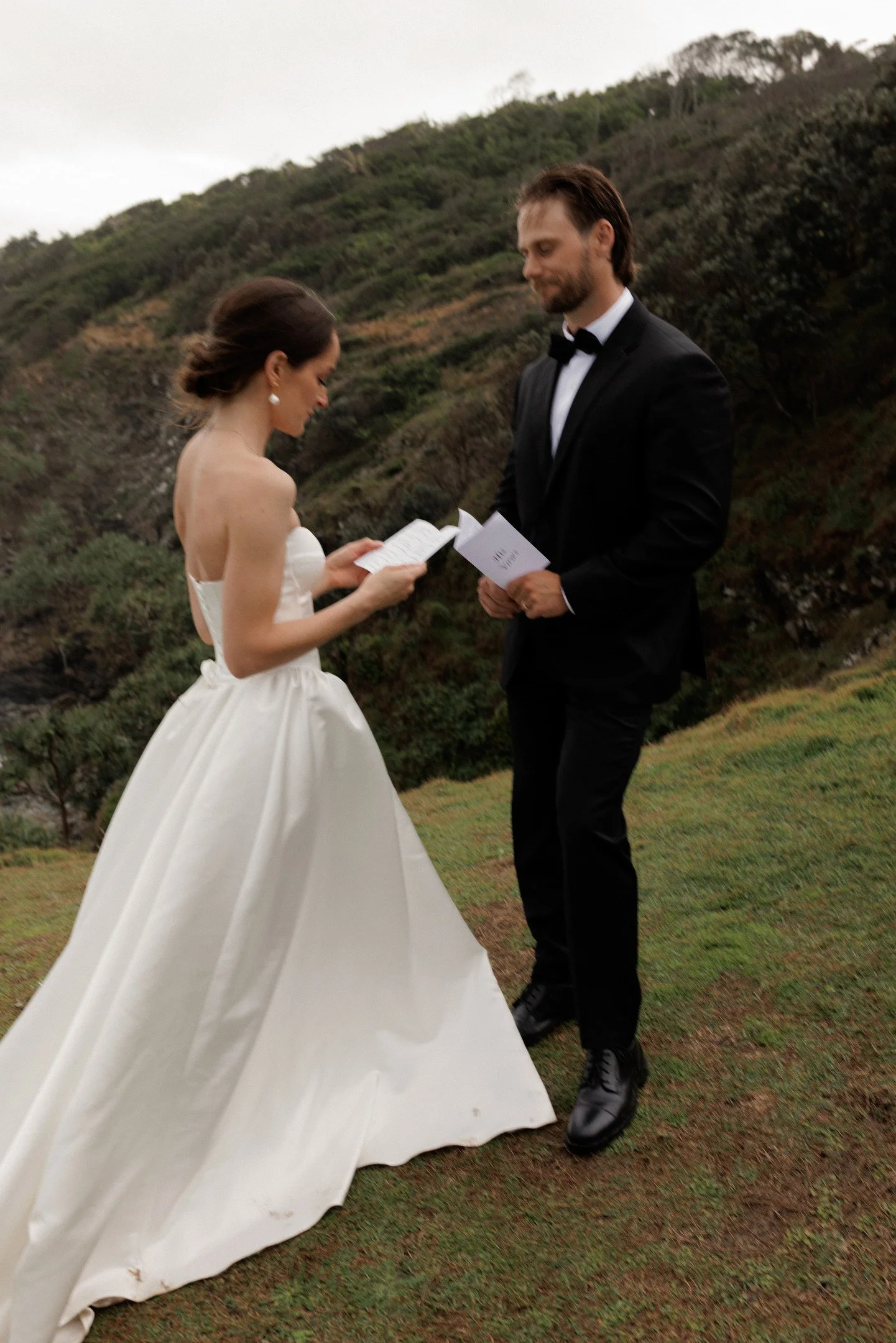 A bride in a white wedding dress and an elegant groom in a black tuxedo standing outdoors on a grassy hillside, reading vows or exchanging rings during a wedding ceremony.