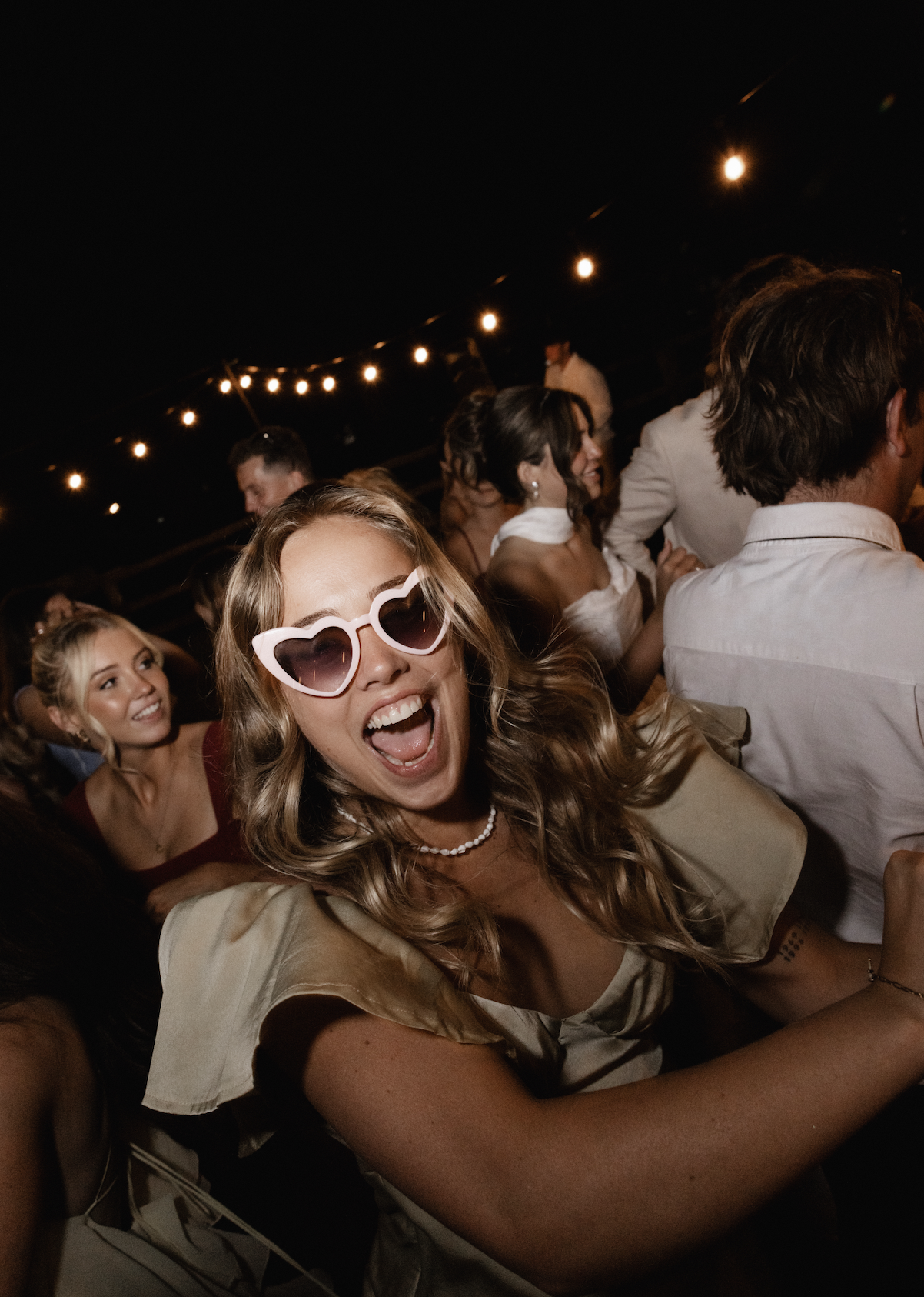 Young woman wearing heart-shaped sunglasses and a pearl necklace smiling and having fun at a party or celebration, with other smiling guests in the background, festive string lights overhead.