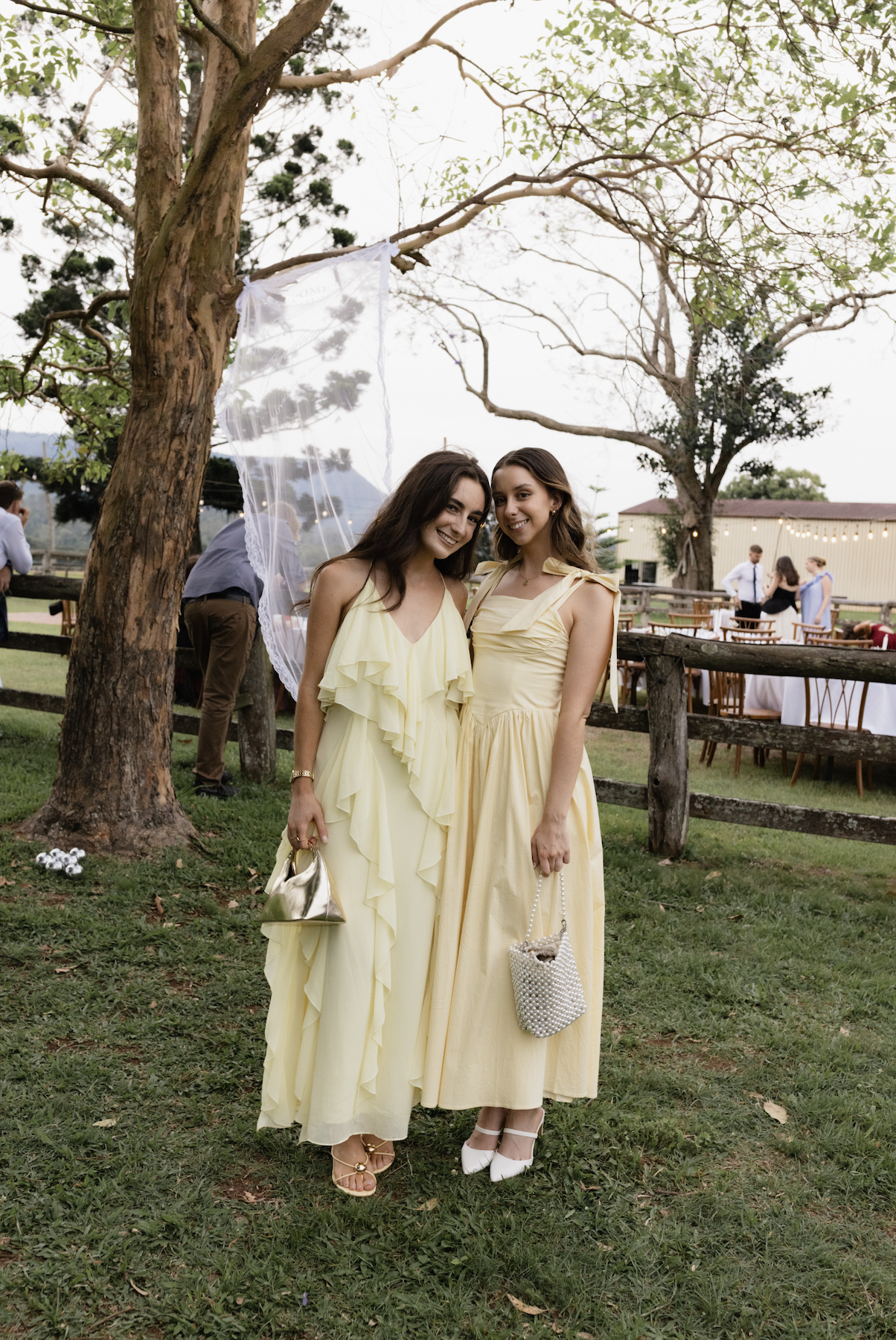 Two women in yellow dresses standing outdoors at a social gathering, smiling at the camera, with trees and a wooden fence in the background.