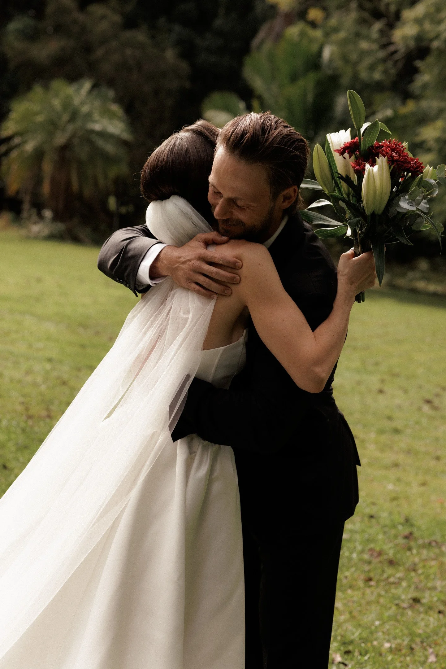 A woman in a white wedding dress hugging a man in a black suit outdoors on a grassy area. The woman is holding a bouquet of white lilies and red flowers. They are smiling and appear happy.