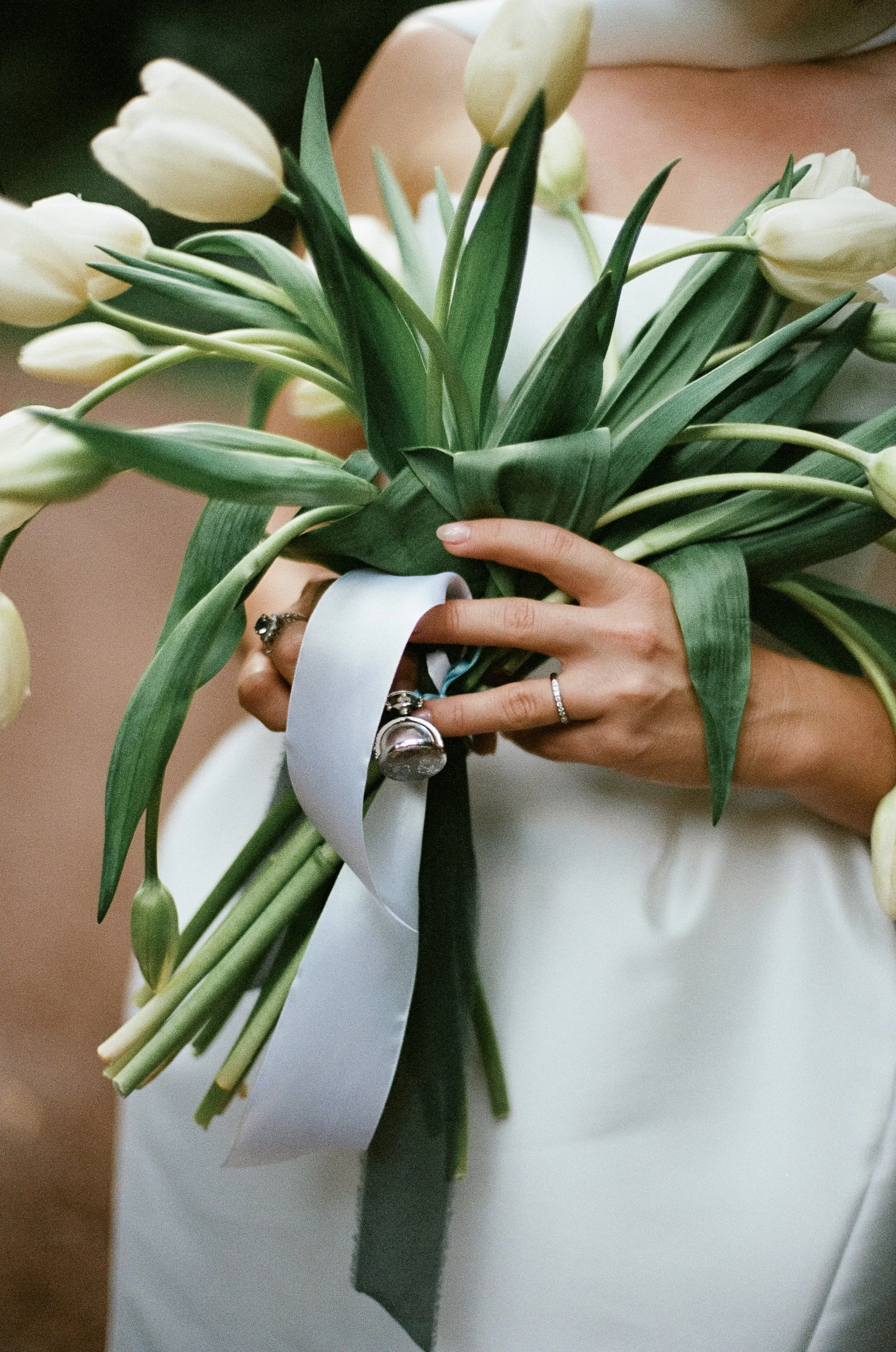 Person holding a bouquet of white tulips with rings on fingers, white ribbon, and green leaves.