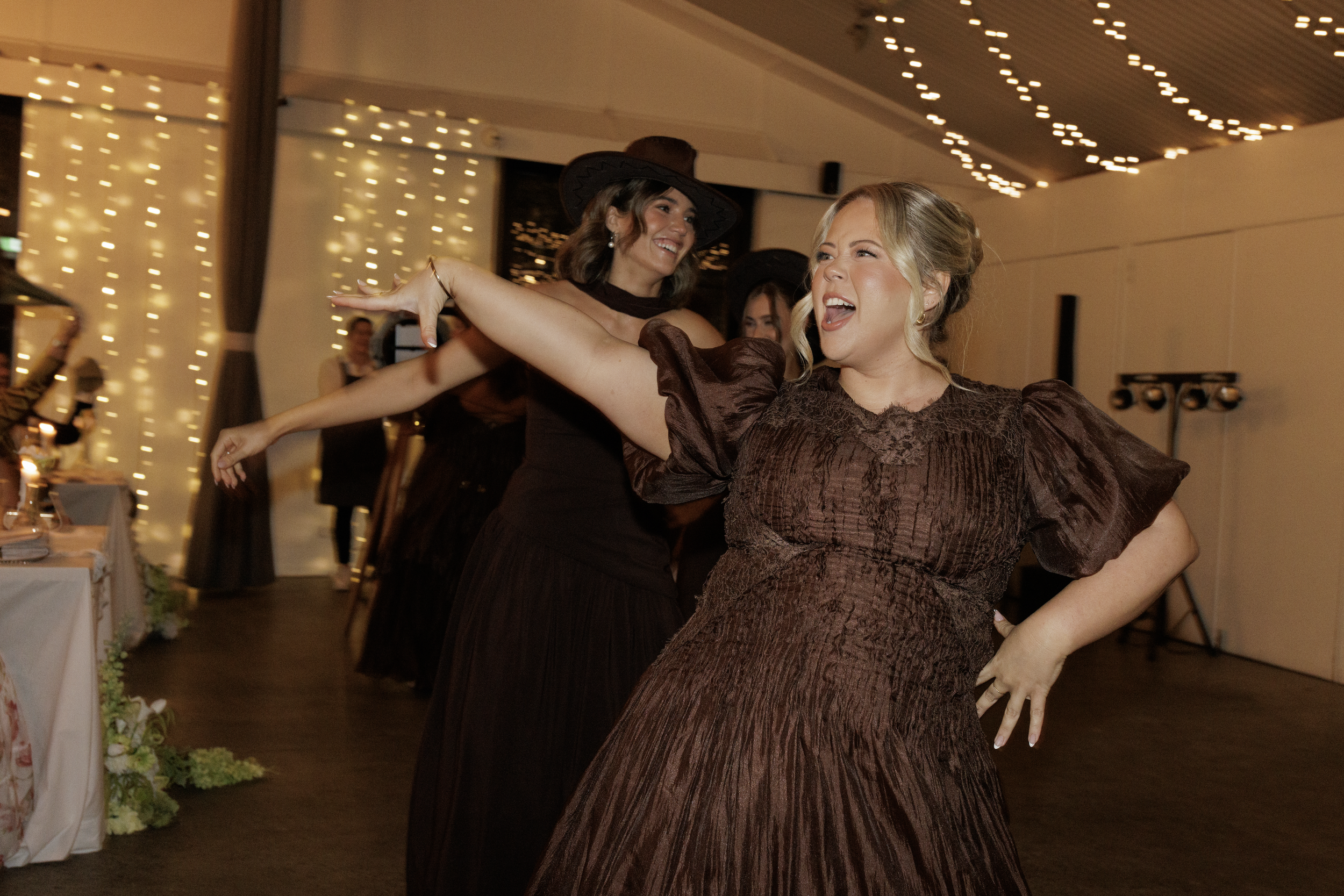 Two women dancing and smiling at a celebration, decorated with string lights and floral arrangements.