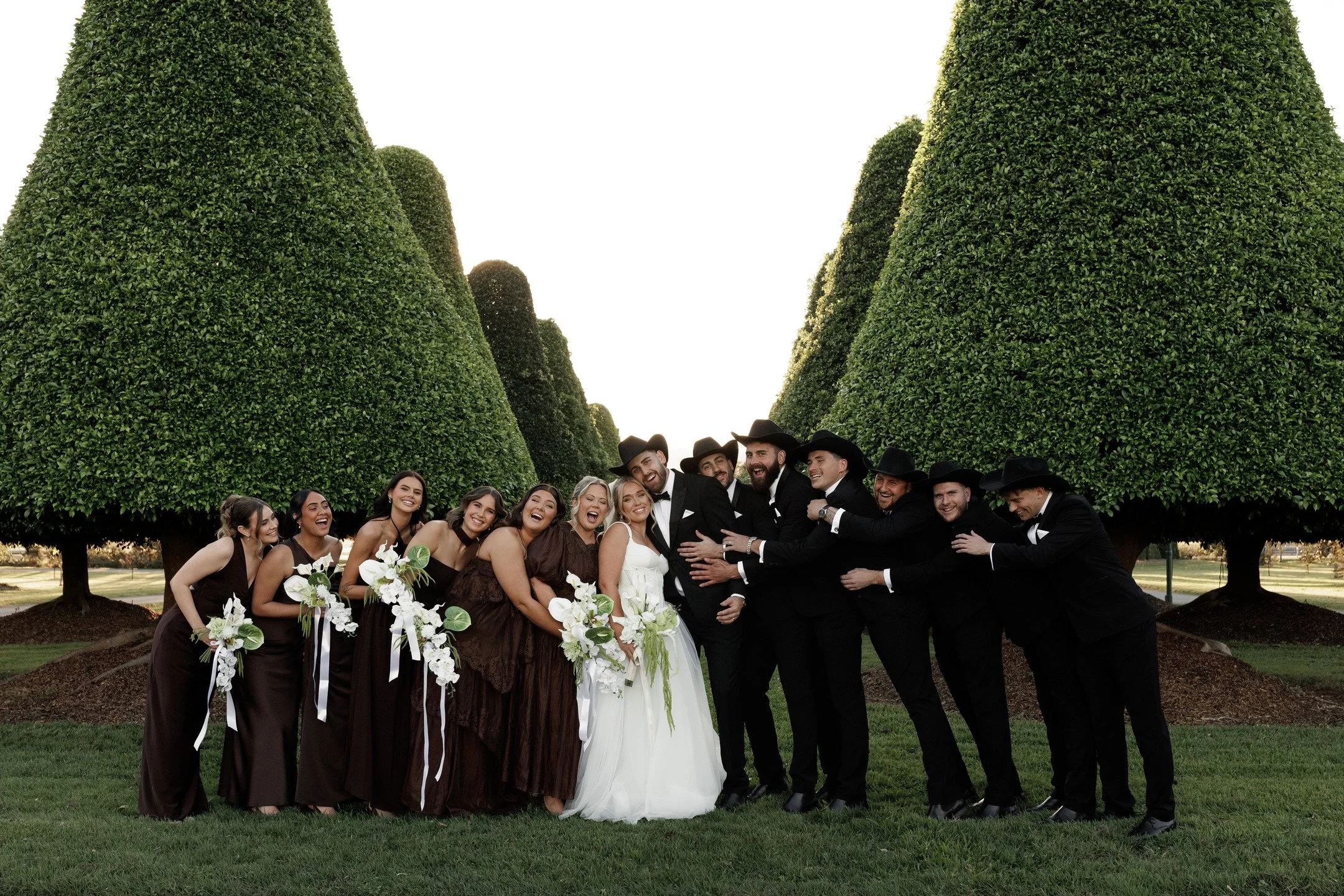 A group of wedding guests in formal attire, standing under decorative topiary trees in a park.