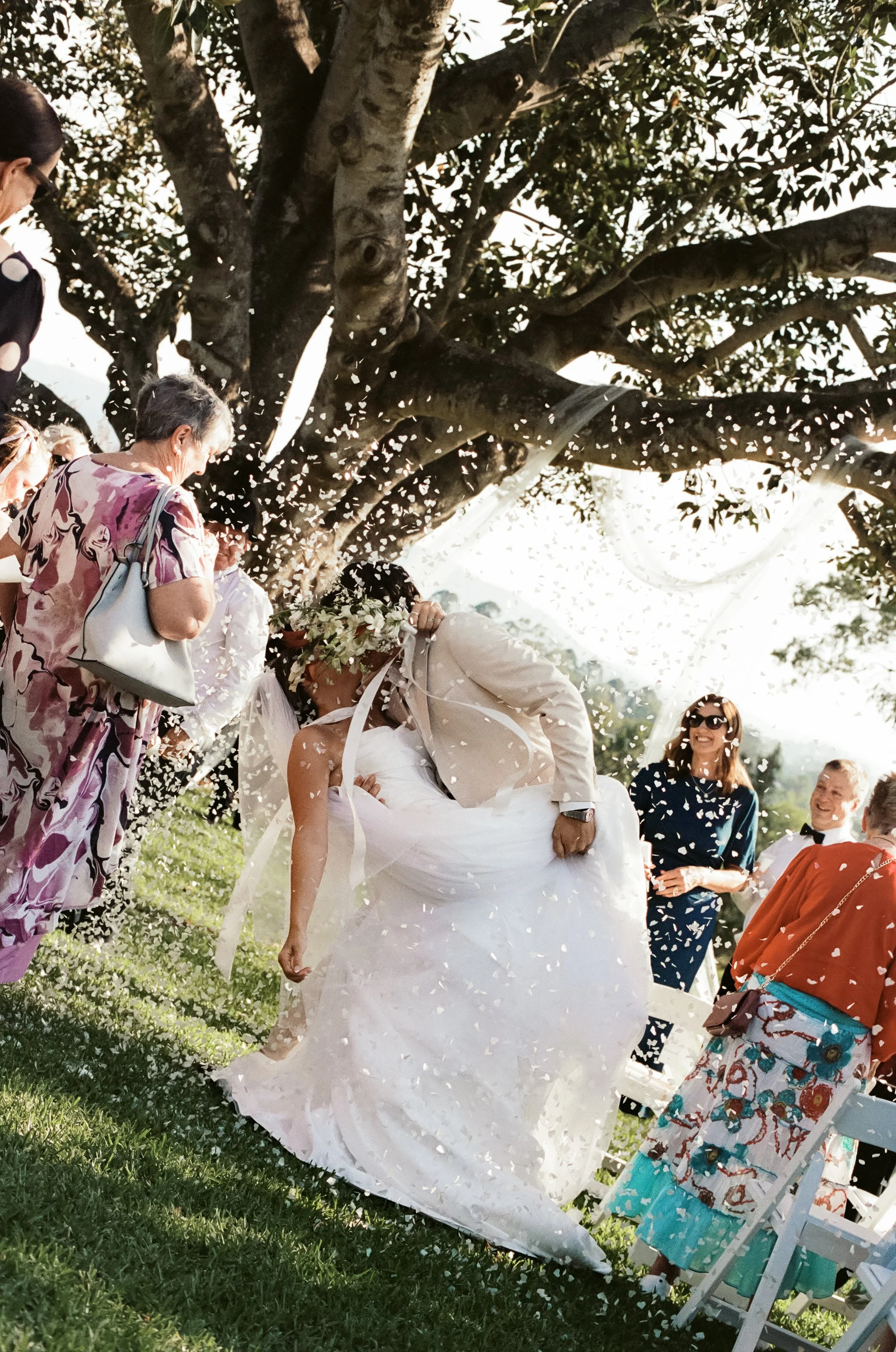 Bride and groom kissing under a tree as guests celebrate and throw flower petals during a wedding ceremony outdoors.