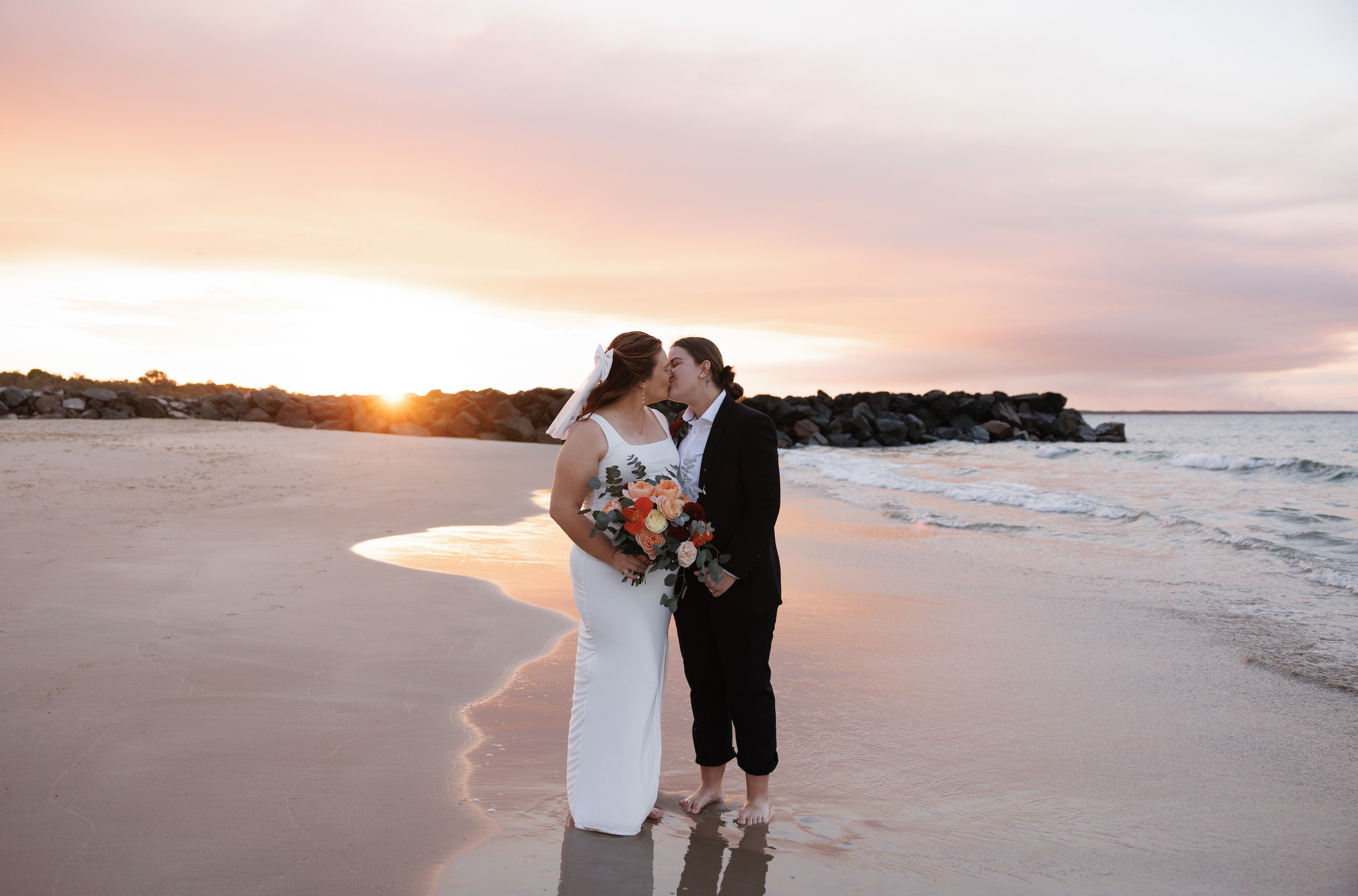 Couple in wedding attire kissing on a beach at sunset, with the bride holding a bouquet of flowers.