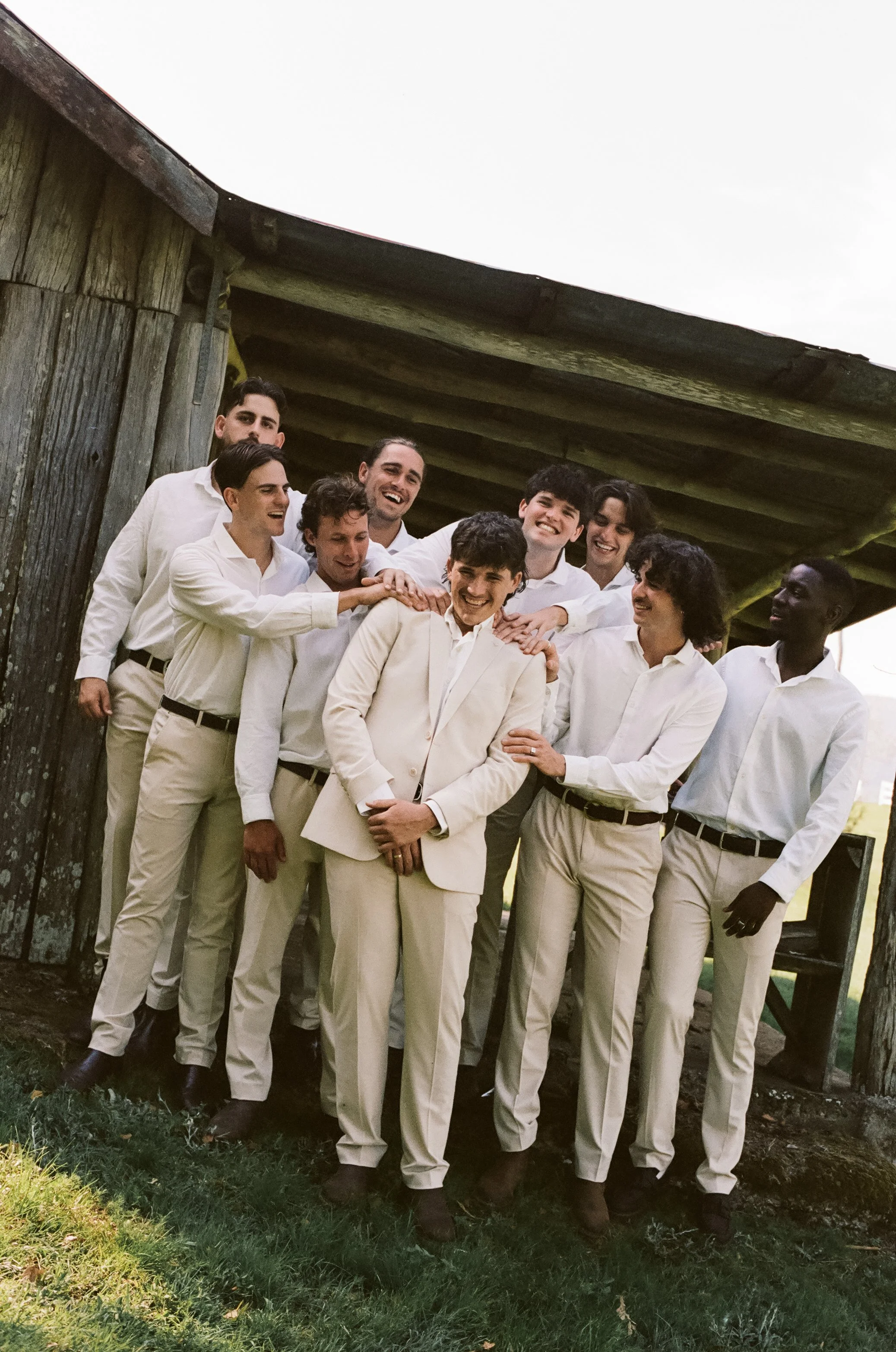 A group of ten men dressed in white shirts and beige pants, gathered together outdoors under a wooden structure, smiling and laughing.