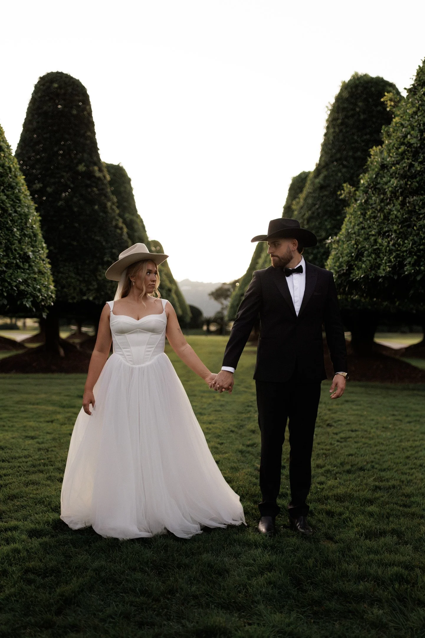 A man and woman dressed in formal attire, holding hands and walking in a garden with trimmed trees and green grass, during sunset or early evening.