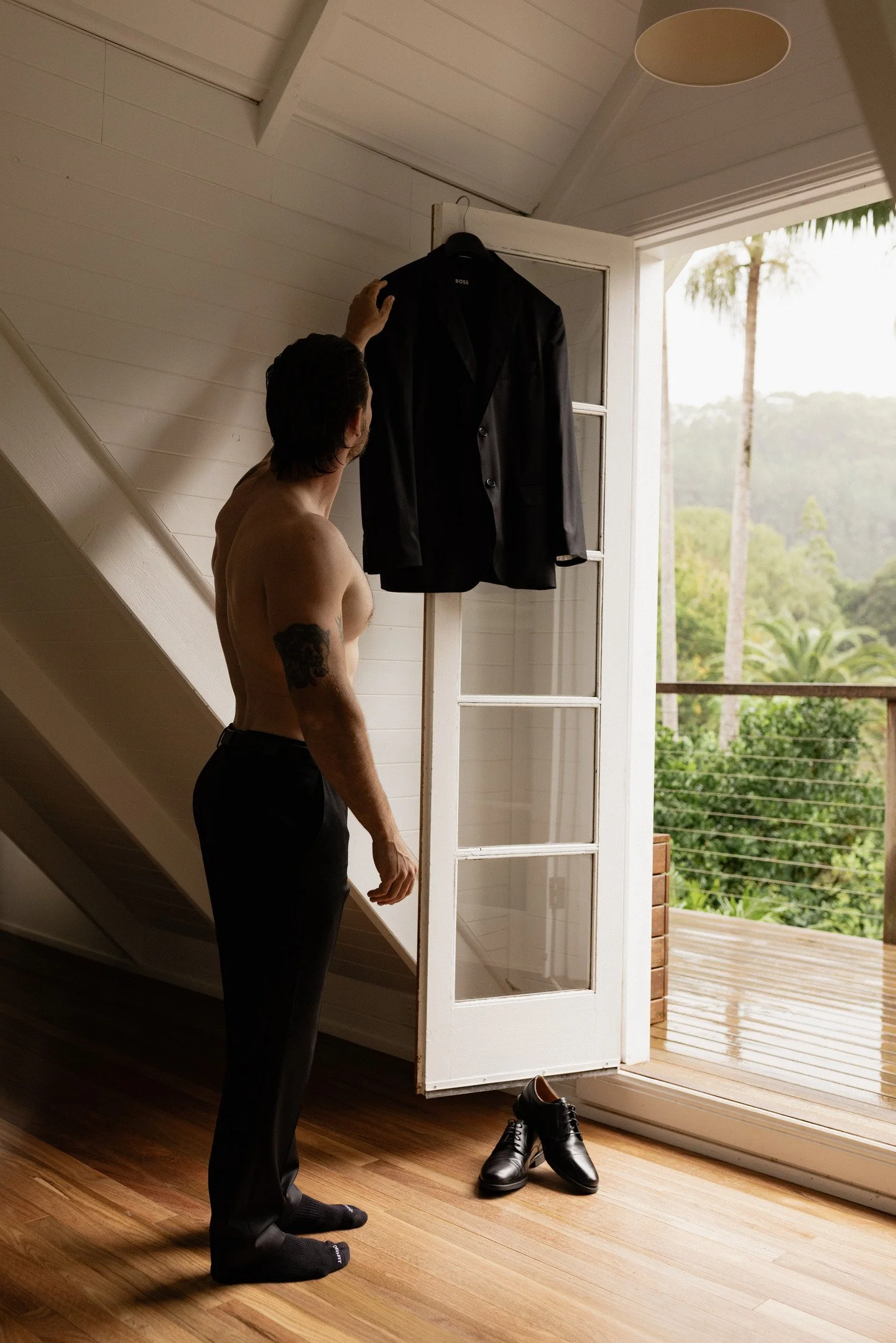 A shirtless man with tattoos on his left arm and wearing socks, standing on a wooden floor inside a room, reaching up to a hanger holding a black suit jacket near an open door leading to a balcony with trees outside.
