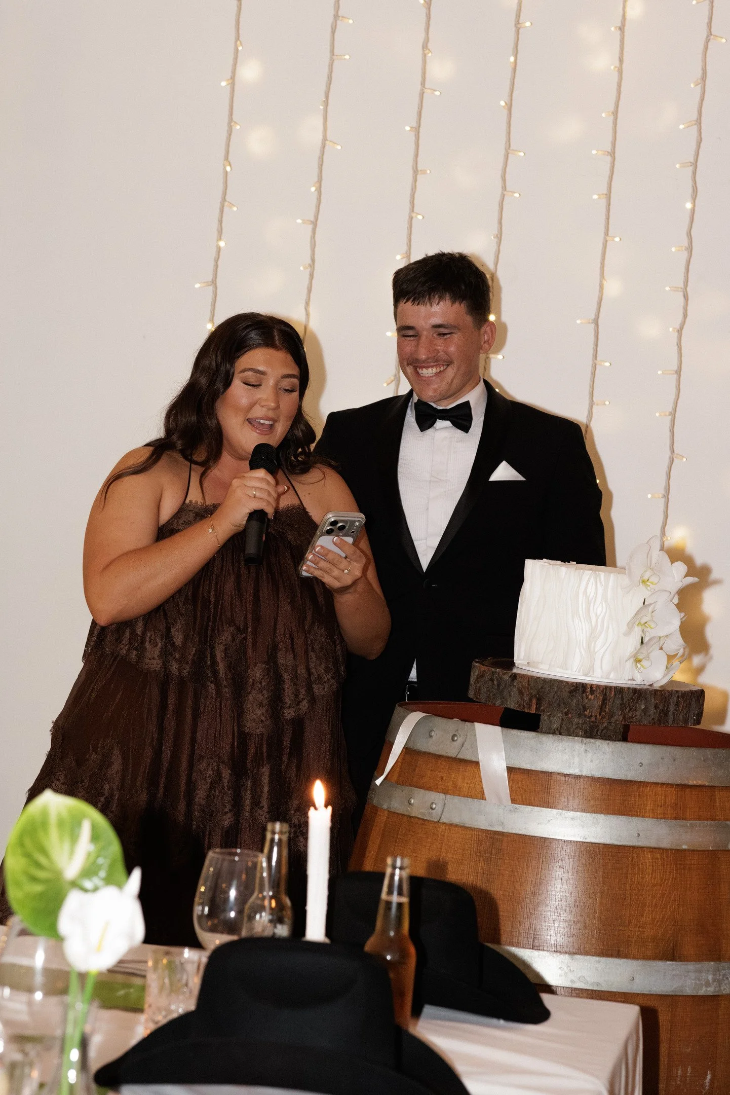 A woman in a brown dress and a man in a black tuxedo with a bowtie are smiling and looking at a phone during a celebration with a white cake decorated with flowers and a candle, set in a decorated venue with string lights in the background.