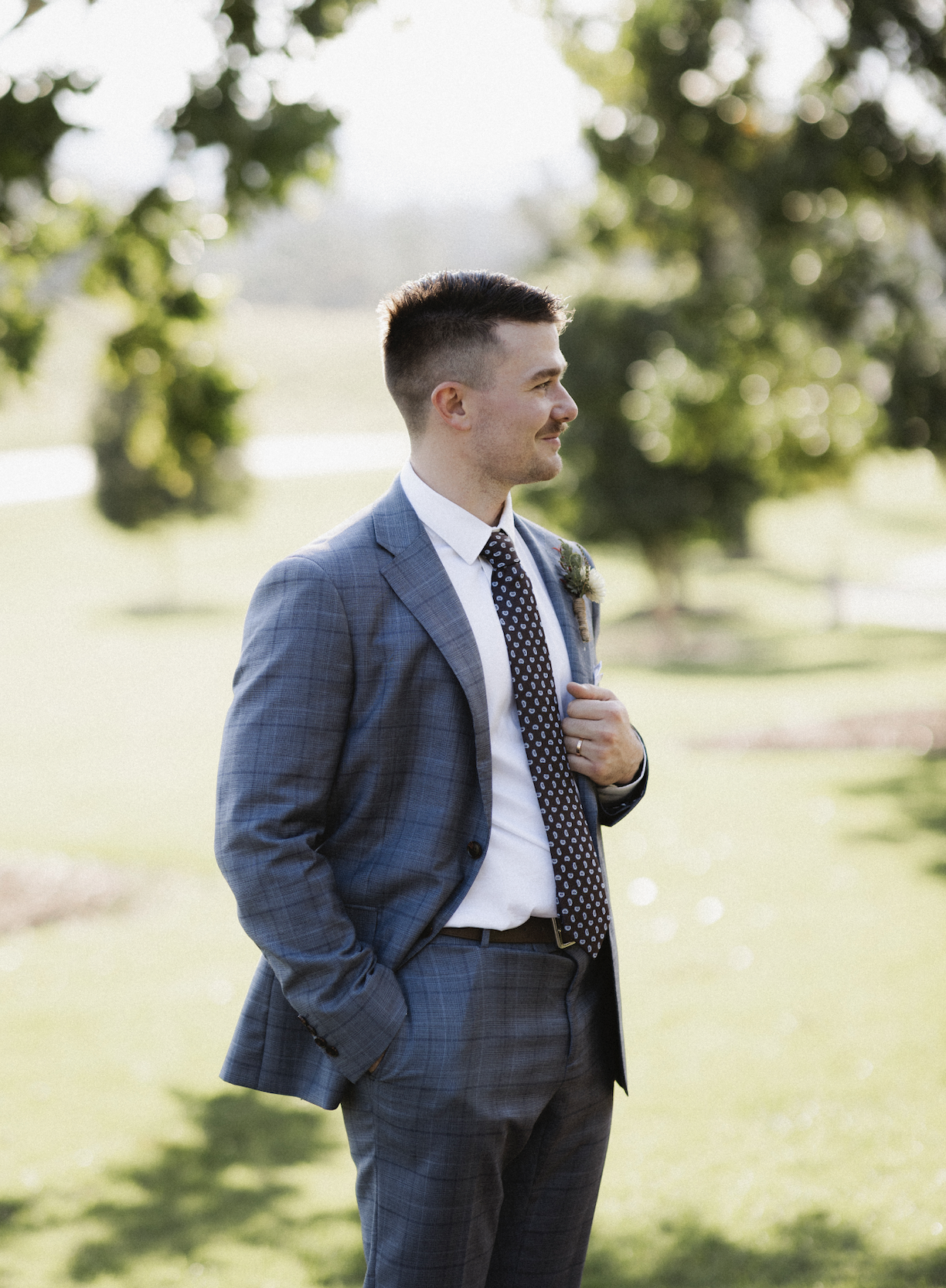 Man in a gray plaid suit, white shirt, and patterned tie, standing outdoors in a park with green grass and trees, smiling sideways, with one hand in his pocket and the other adjusting his jacket.