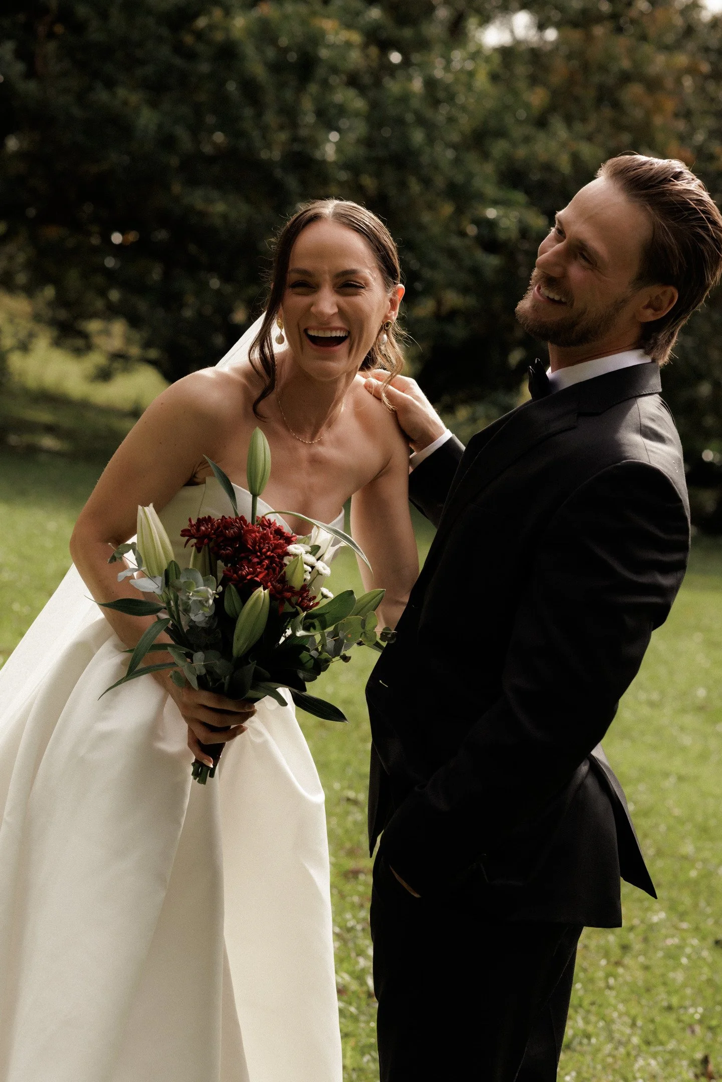 Smiling bride in a white wedding gown holding a bouquet of flowers, laughing and looking at groom who is dressed in a black tuxedo, outdoors on a green lawn with trees in the background.
