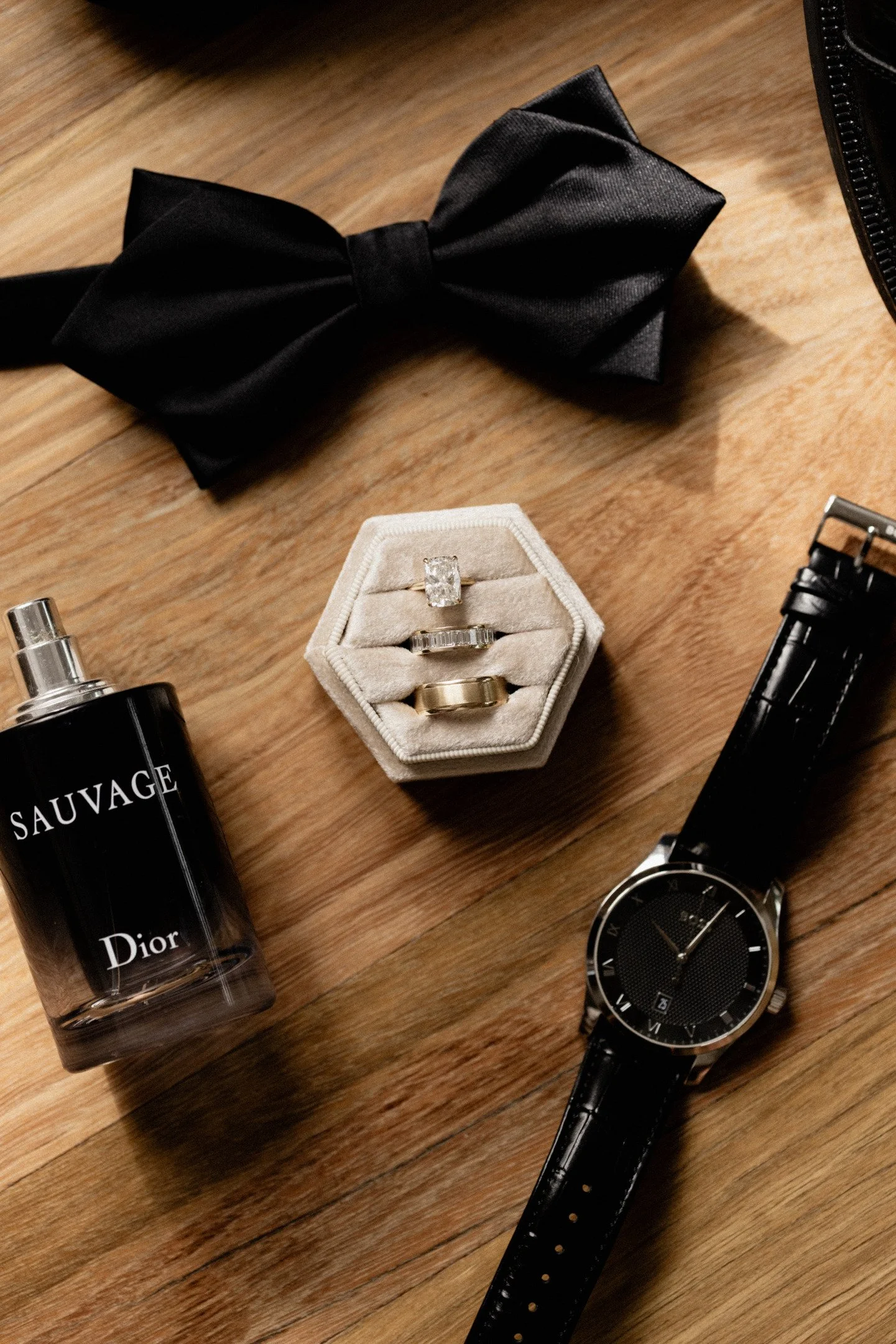 Black bow tie, a diamond ring in a beige ring box, a silver watch with a black face, Dior Sauvage perfume, and a black watch on a wooden surface.