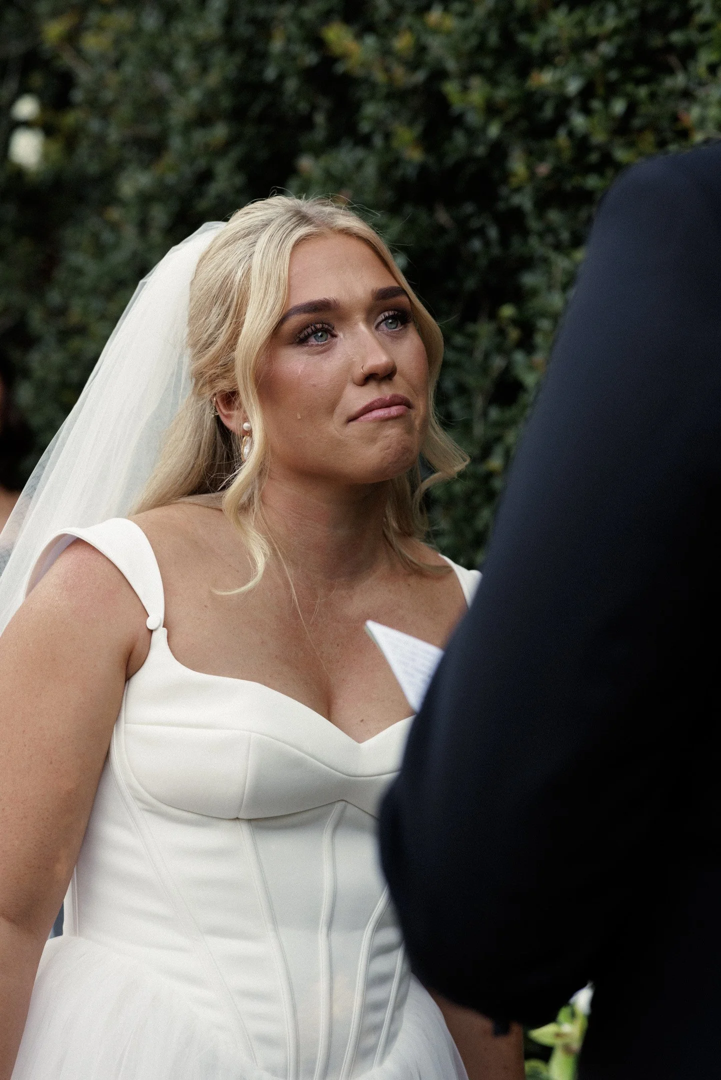 A bride with blonde hair wearing a white wedding dress and veil, standing outdoors during her wedding ceremony, visibly emotional with tears in her eyes, facing a man in a dark suit.