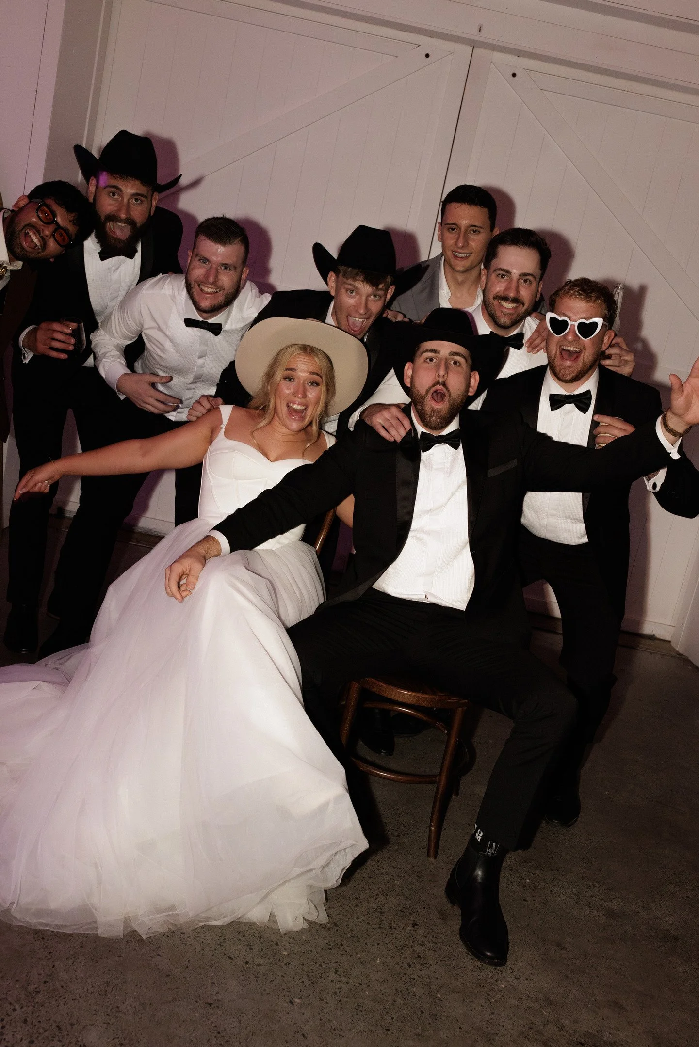 Group of people celebrating at a wedding reception. The bride is in a white gown, and the groom is in a tuxedo. Others are dressed in formal attire, some with hats and fun accessories like heart-shaped sunglasses, smiling and posing for the photo.