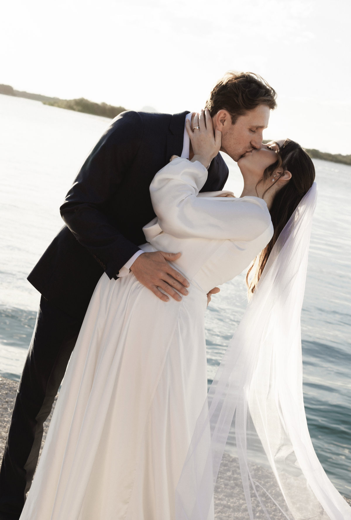 A bride and groom kissing by a body of water during sunset, with a blurred shoreline in the background.