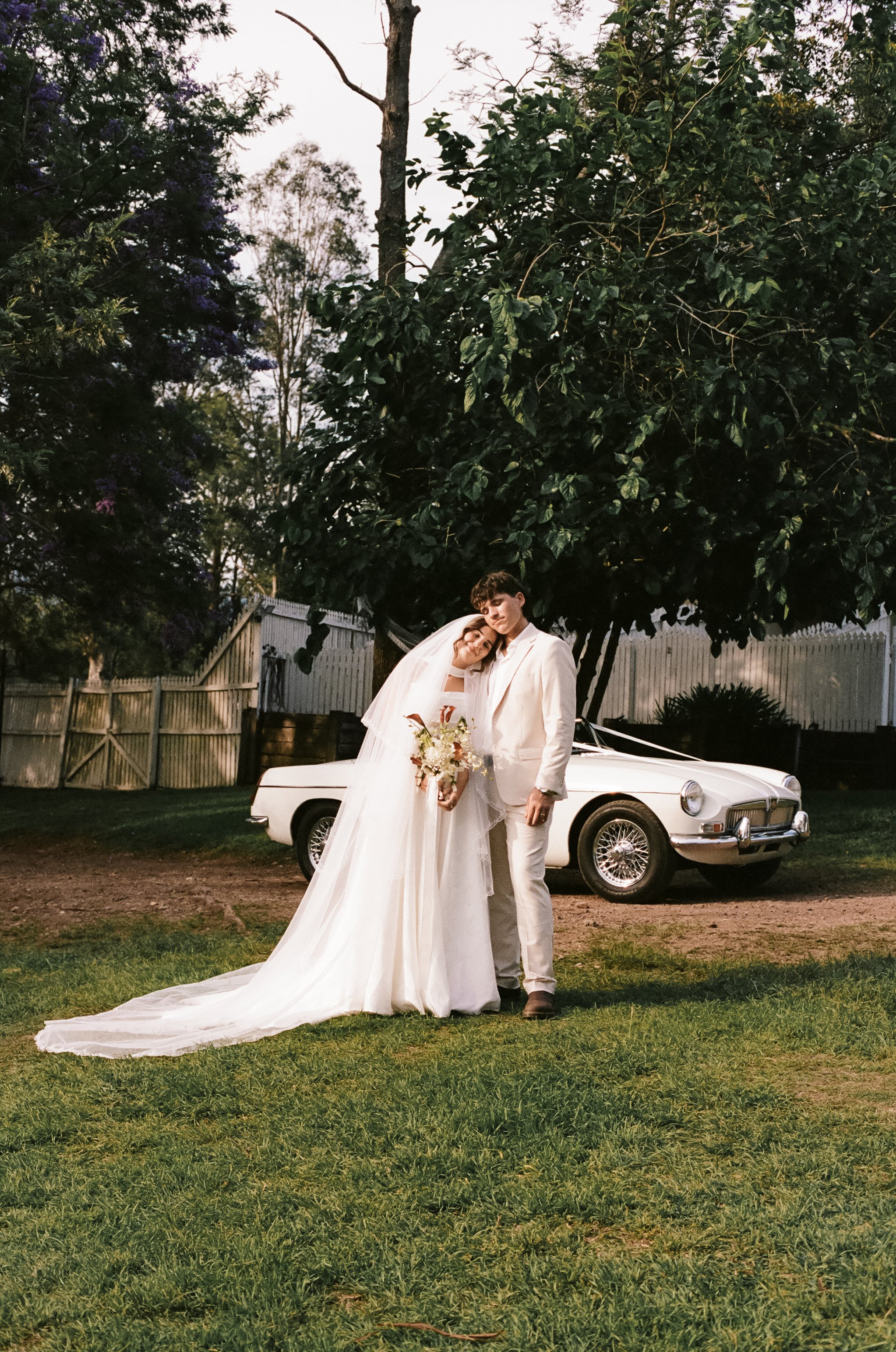 A bride and groom dressed in wedding attire standing on grass in front of a vintage white car and large trees during dusk, with the bride leaning her head on the groom's shoulder and holding a bouquet.