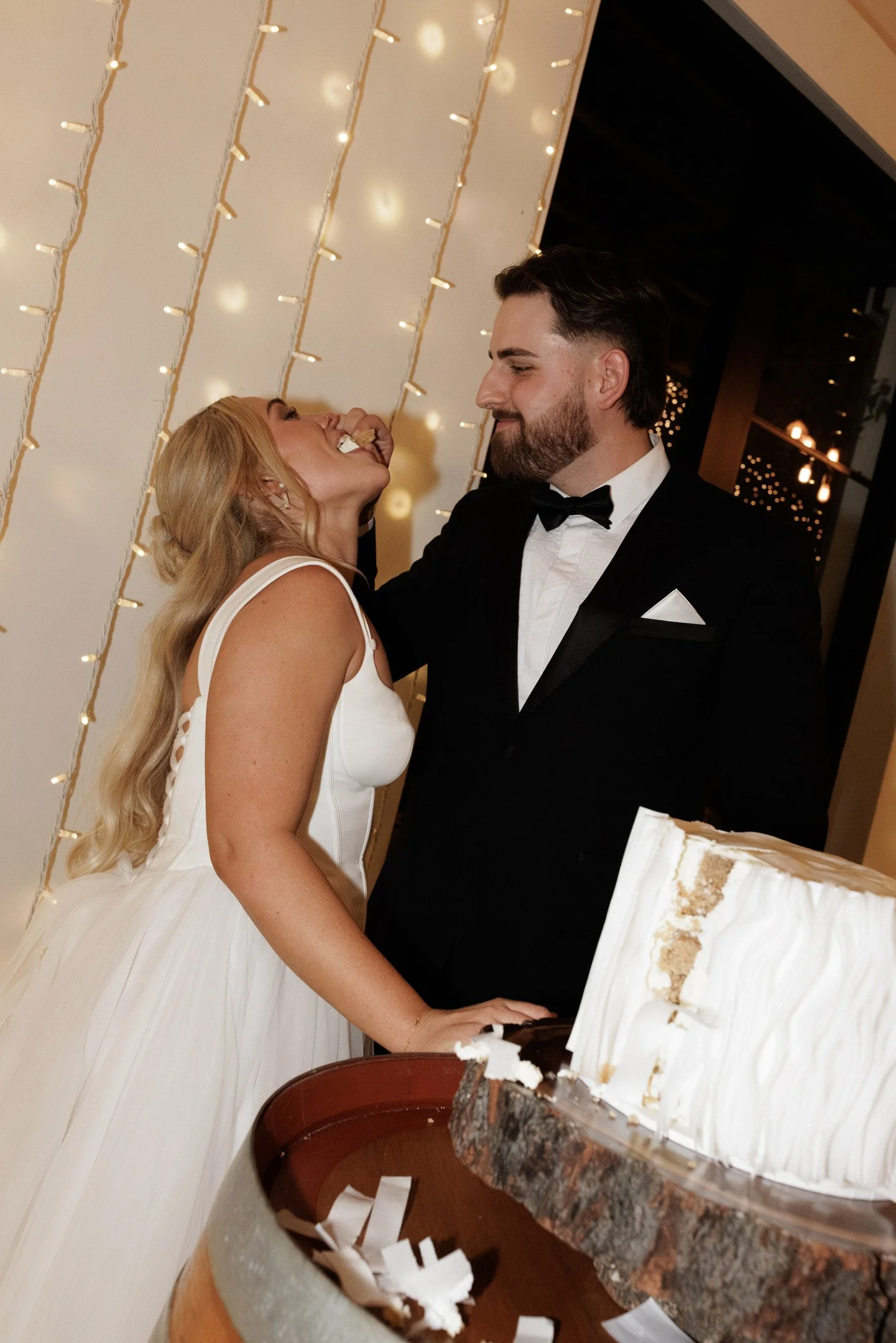 A bride and groom dancing at a wedding reception with a decorated backdrop of string lights. The bride is wearing a white wedding gown, and the groom is in a black tuxedo. A cake is partially visible in the foreground.