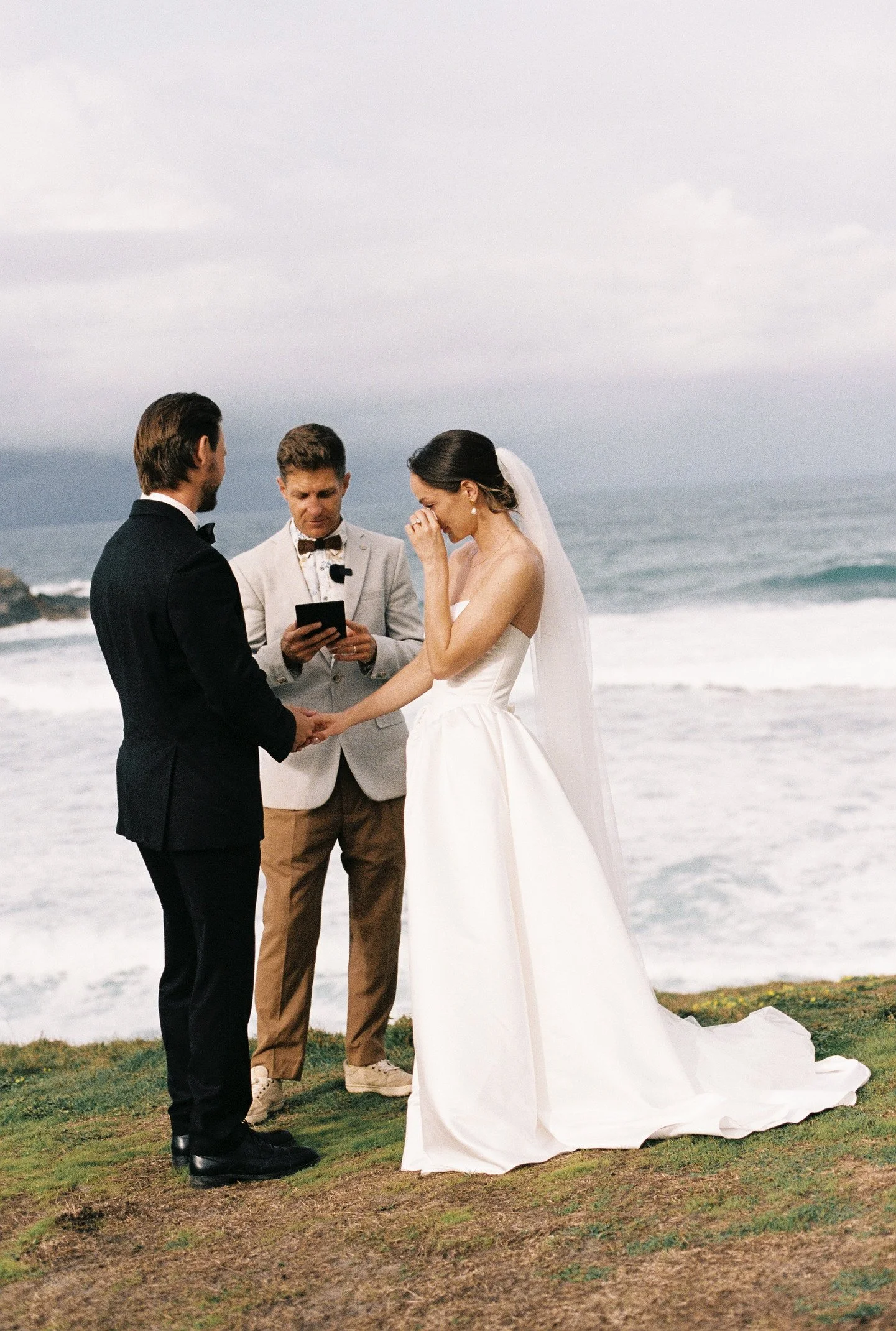 A wedding ceremony taking place outdoors on a grassy area near the ocean, with a bride and groom holding hands and facing each other, an officiant standing between them, and the bride in a white wedding dress and veil, while the groom is in a black t